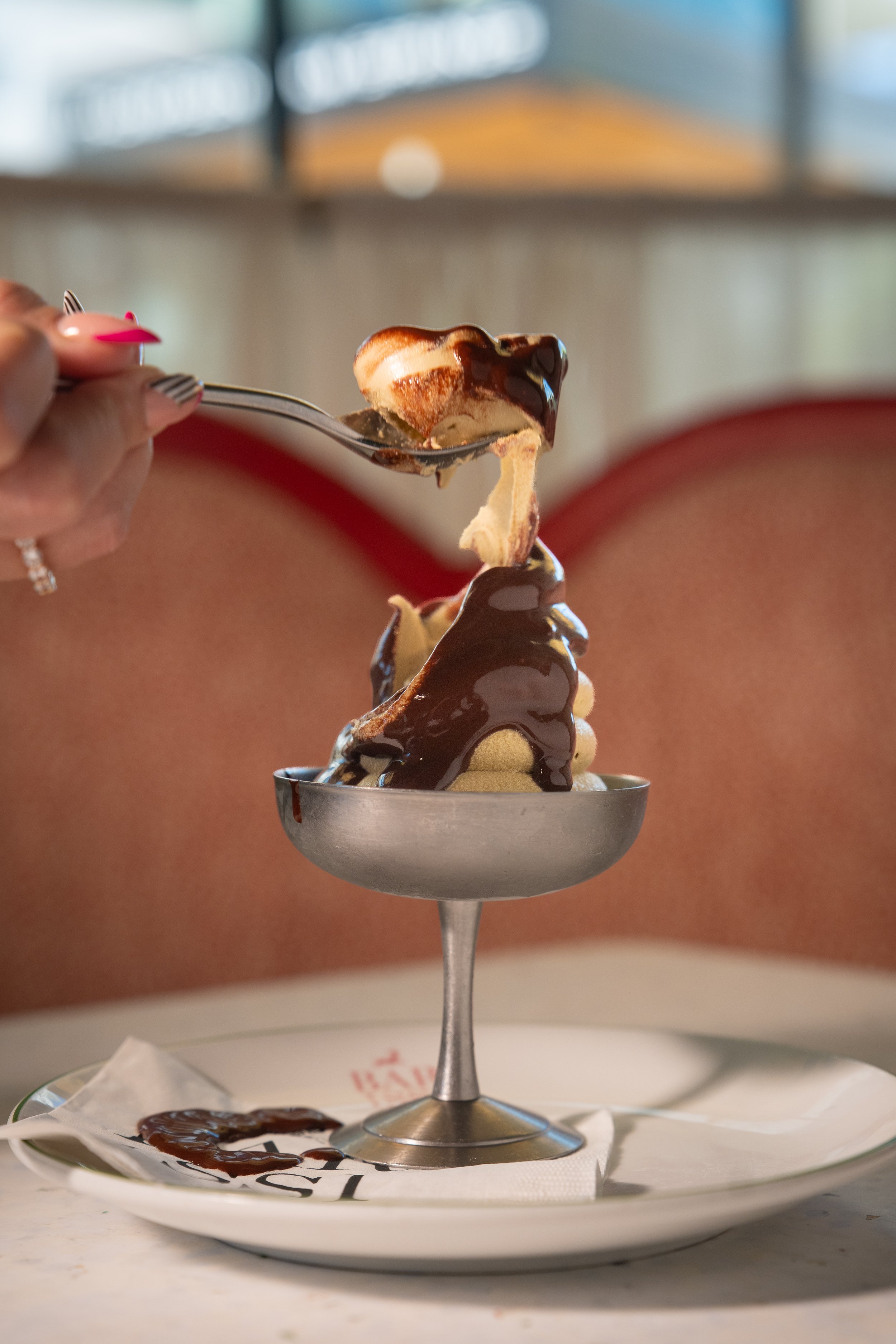 A dessert with ice cream topped with chocolate sauce on a metal stand, placed on a white plate with a napkin underneath.