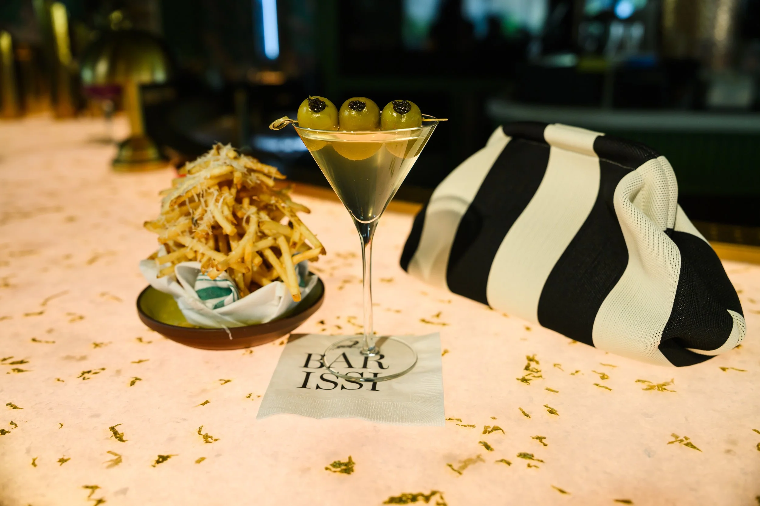 A martini glass with three green olives stuffed with black olives on a pick, sitting on a table at a bar or restaurant. To the left, there is a basket of cooked fries, and to the right, a black and white striped cloth bag. The background appears to b