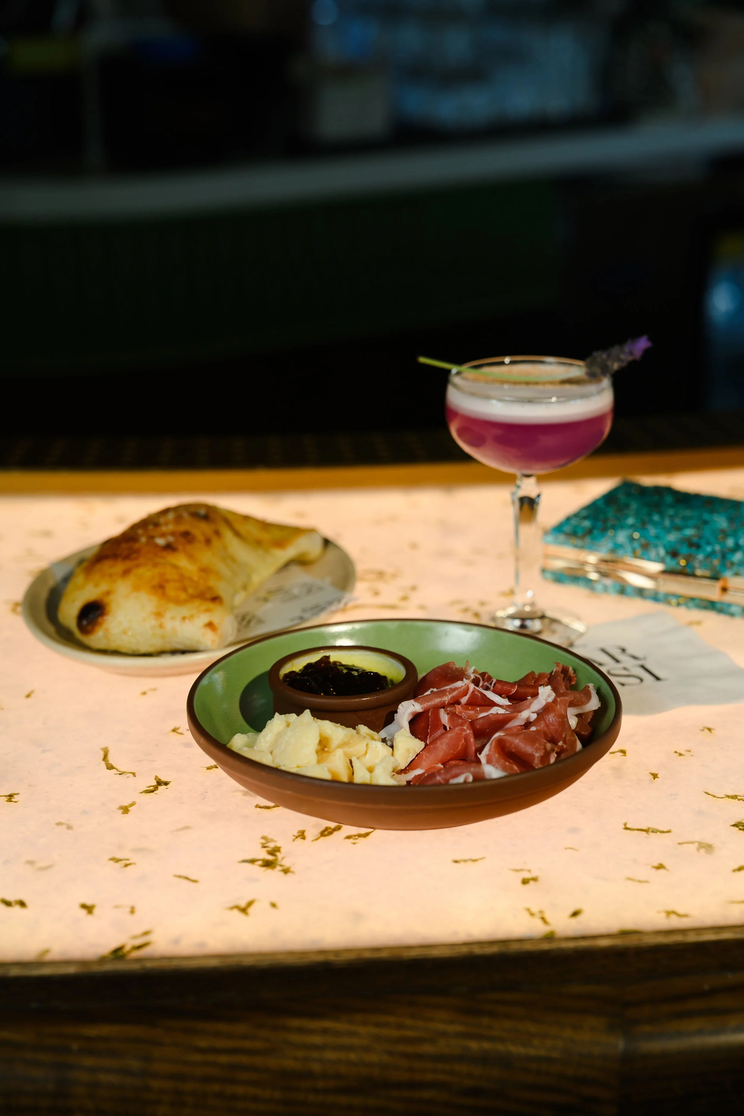 A plate of cheese, cured meats, and a small dish of jam or preserves on a wooden table. In the background, a glass of pink cocktail with a garnish and a croissant.