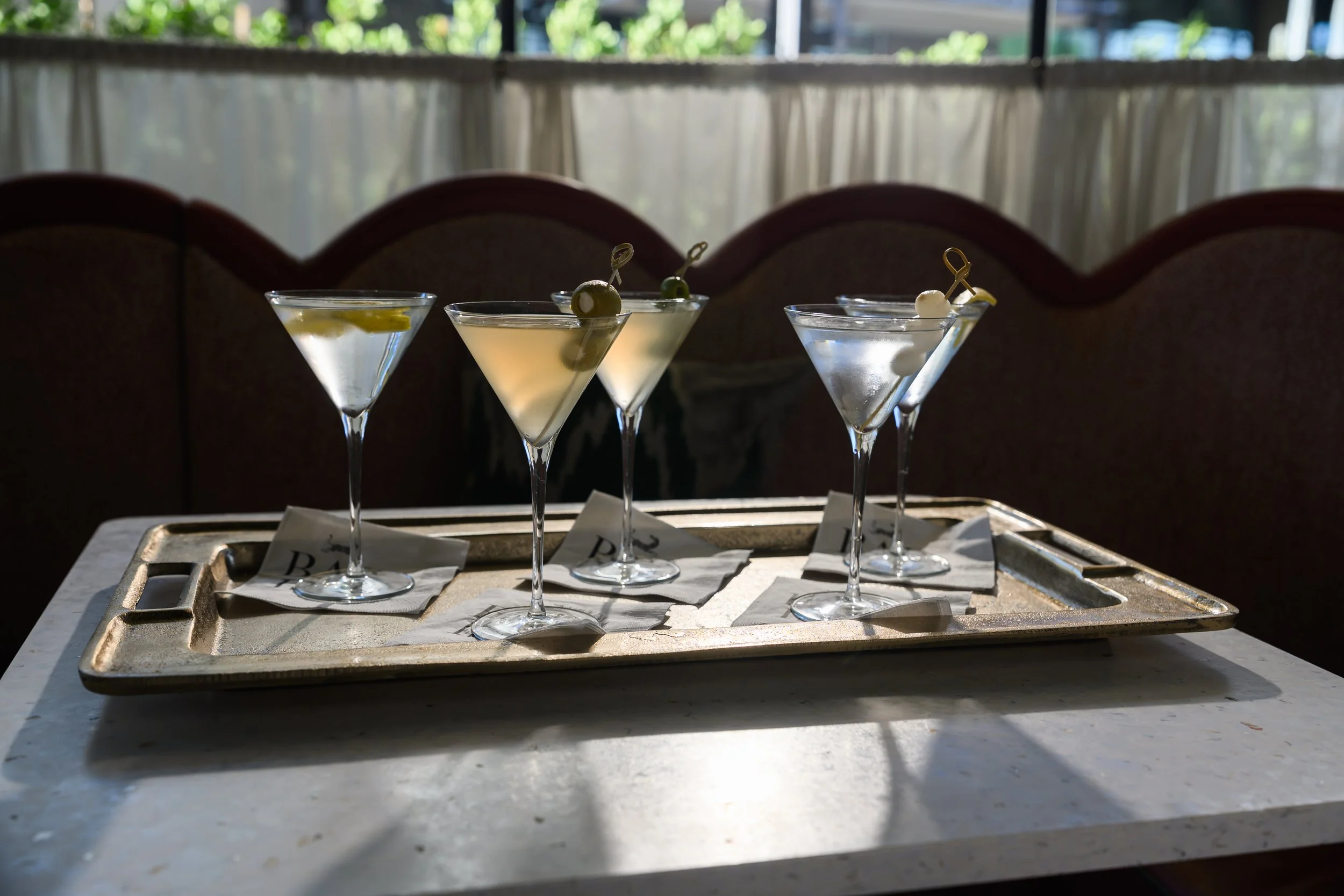 Six cocktail glasses with olives and lemon slices on a tray, placed on a white table in a brightly lit room with natural light coming through the windows.