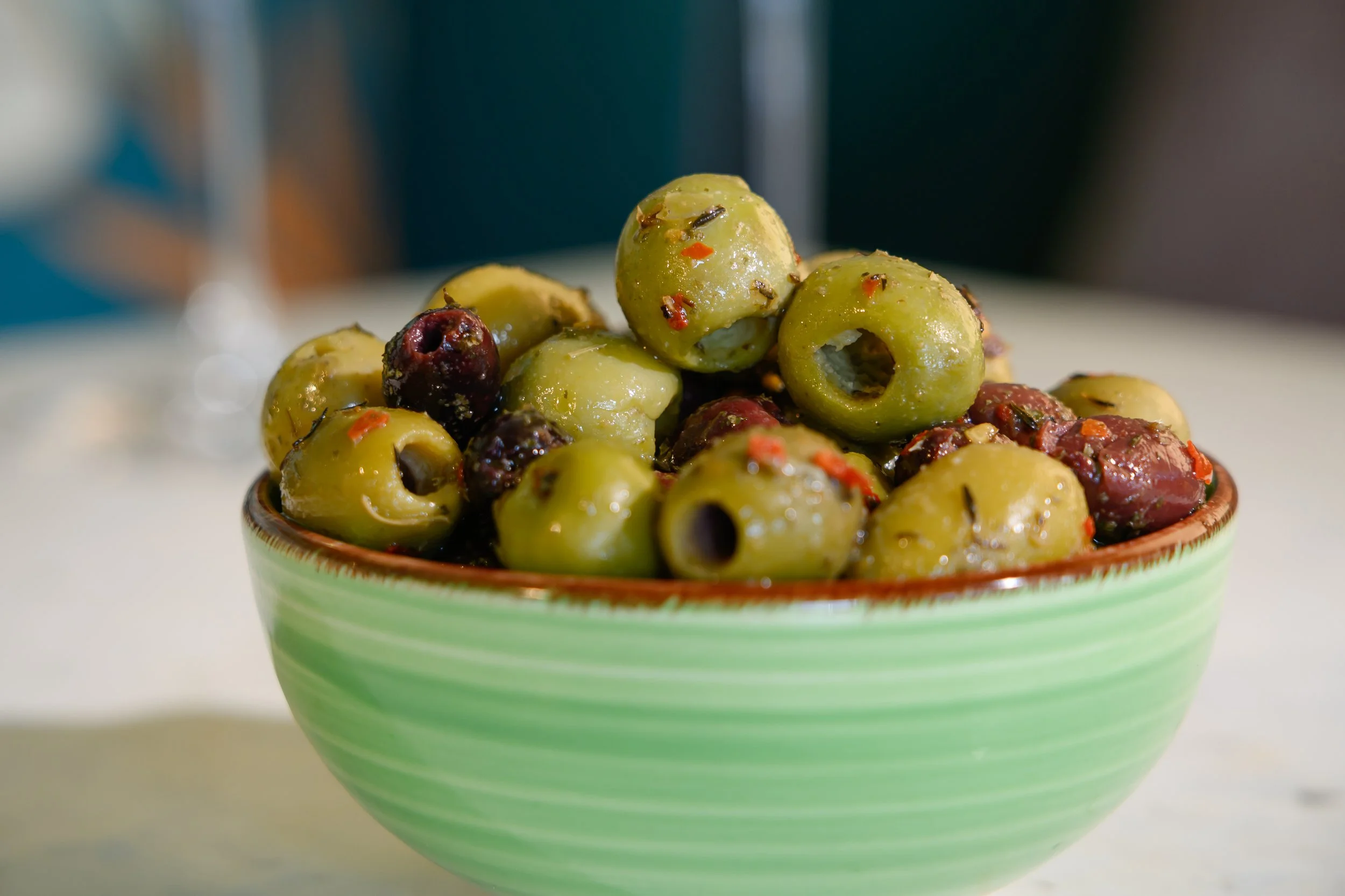 A bowl of green olives with red pepper flakes and herbs on a white surface.