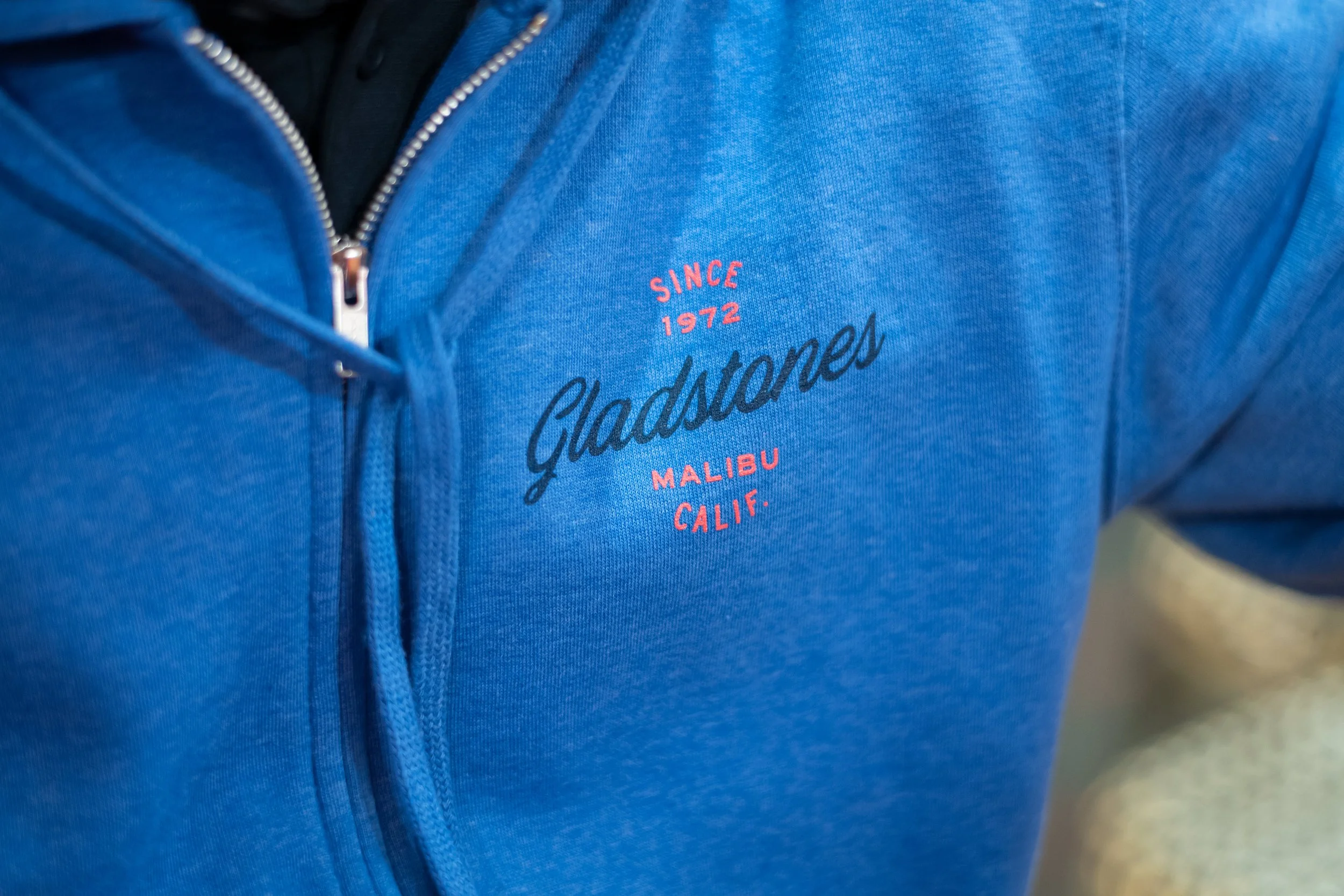 Close-up of a person wearing a blue zip-up jacket with a small embroidered design that reads 'Since 1972 Malibu Calif.'