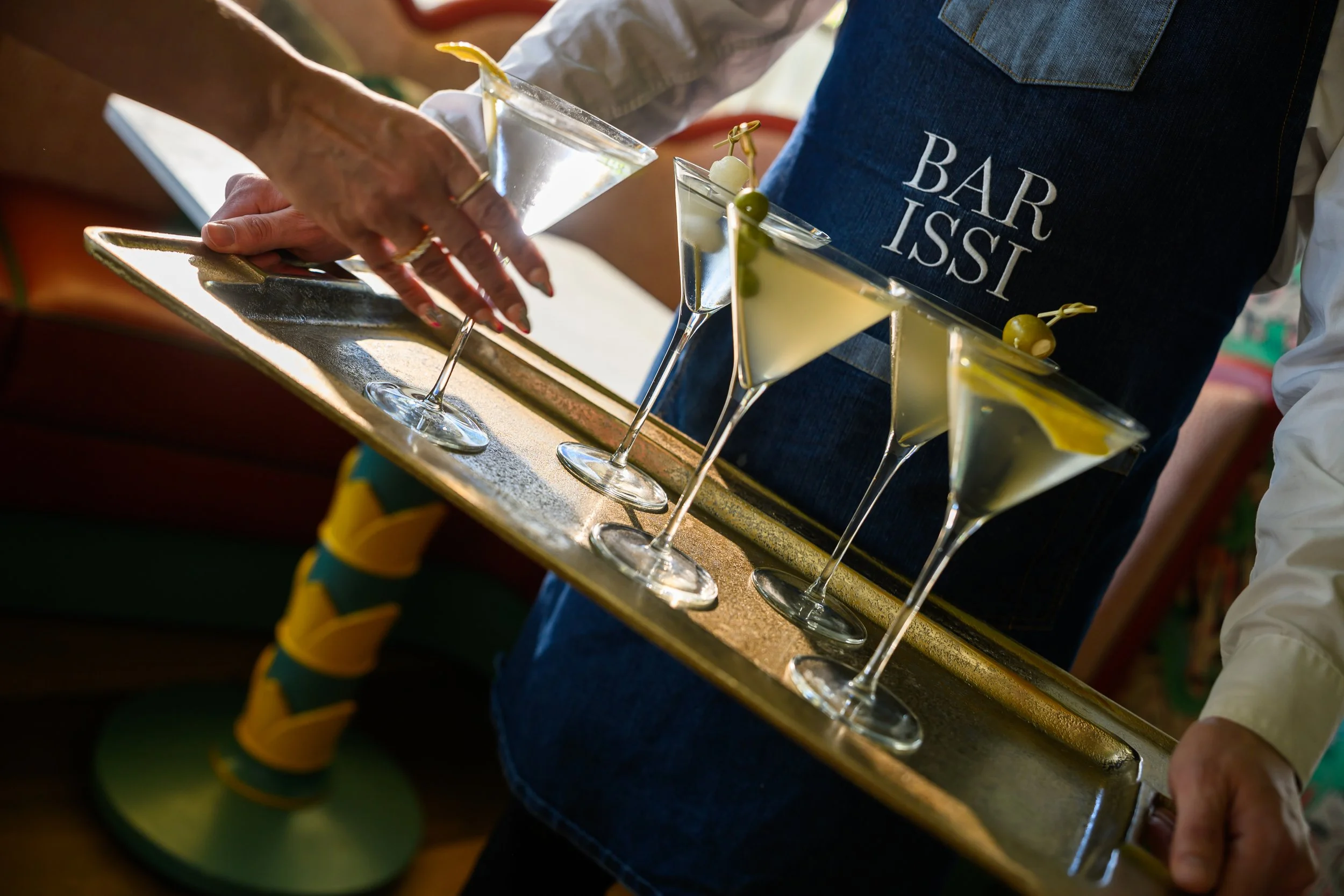 Close-up of a bartender's hand preparing cocktails at a bar, with four martini glasses filled with a yellow beverage garnished with green olives, on a wooden bar counter.