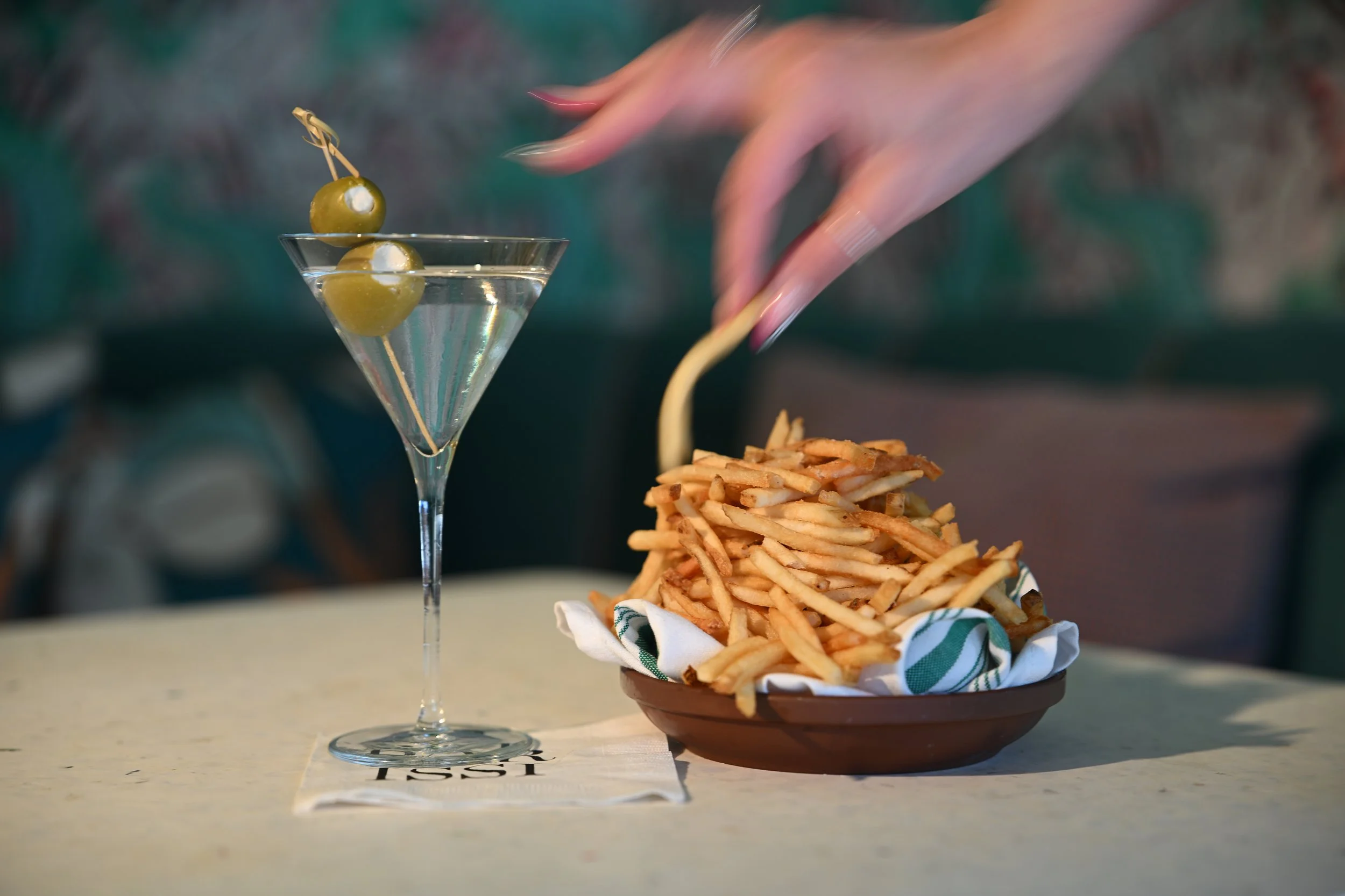 A martini glass with water and green olives on a skewer, next to a basket of French fries on a table.