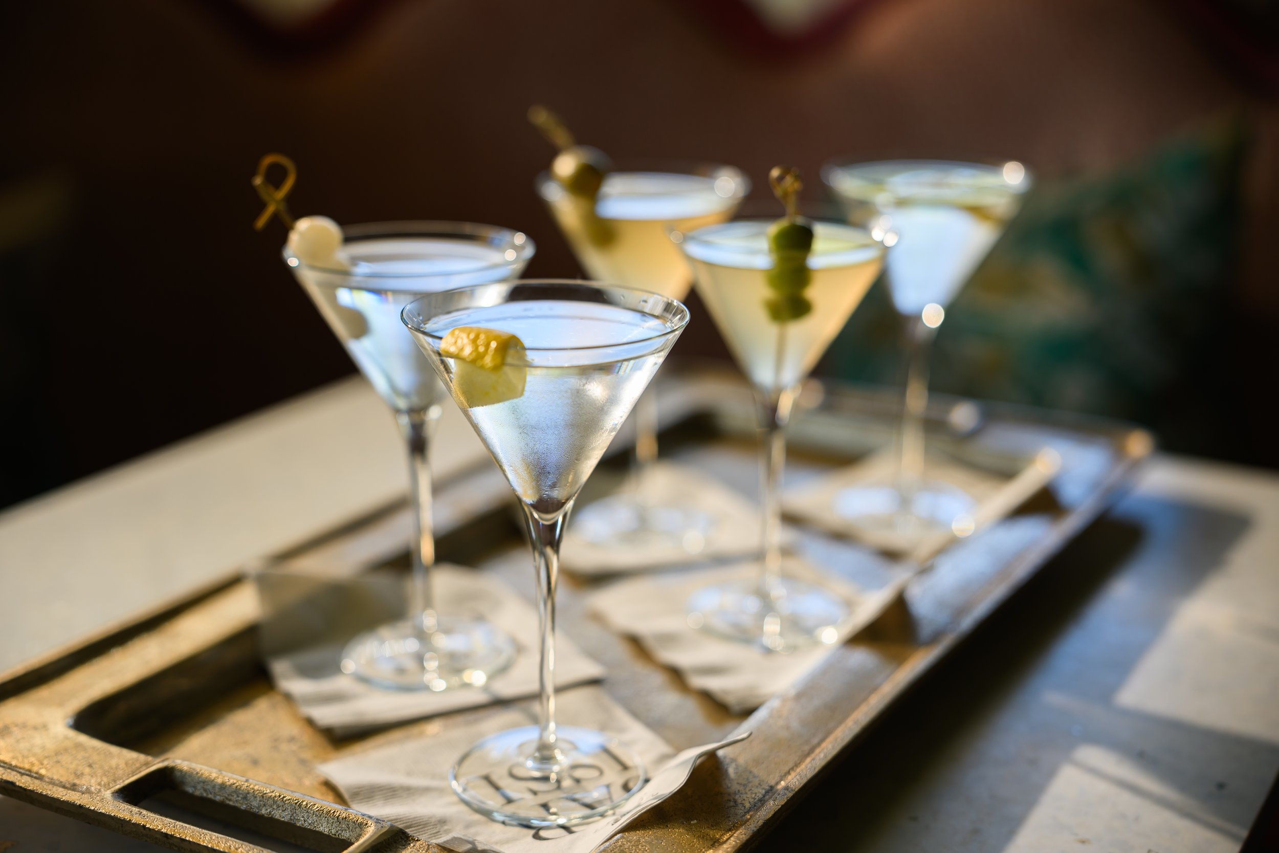 Five elegant martini glasses filled with a clear cocktail, garnished with olives, set on a rustic wooden tray.
