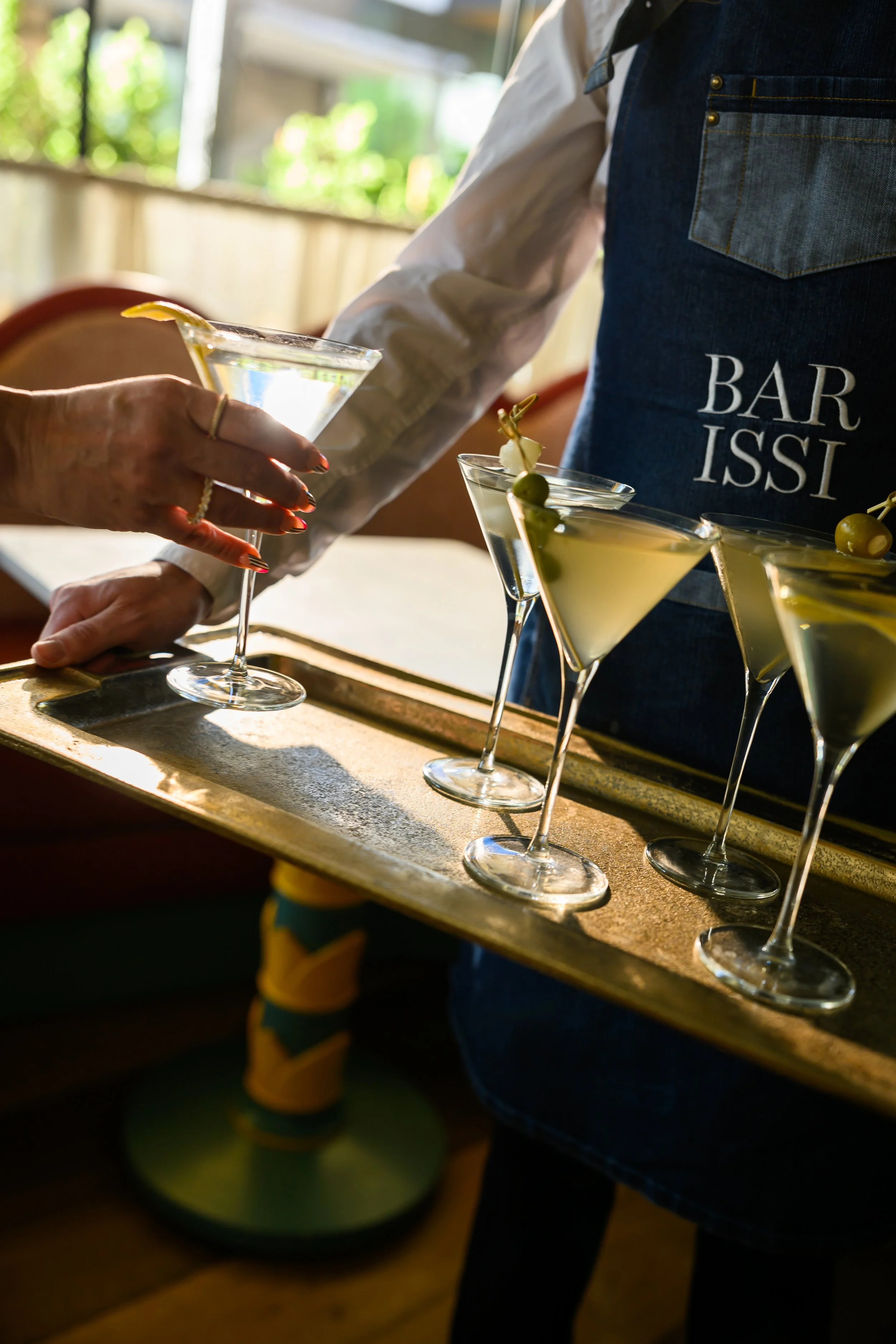 Bartender in a denim apron serving martinis with olives garnished on a tray of five drinks.