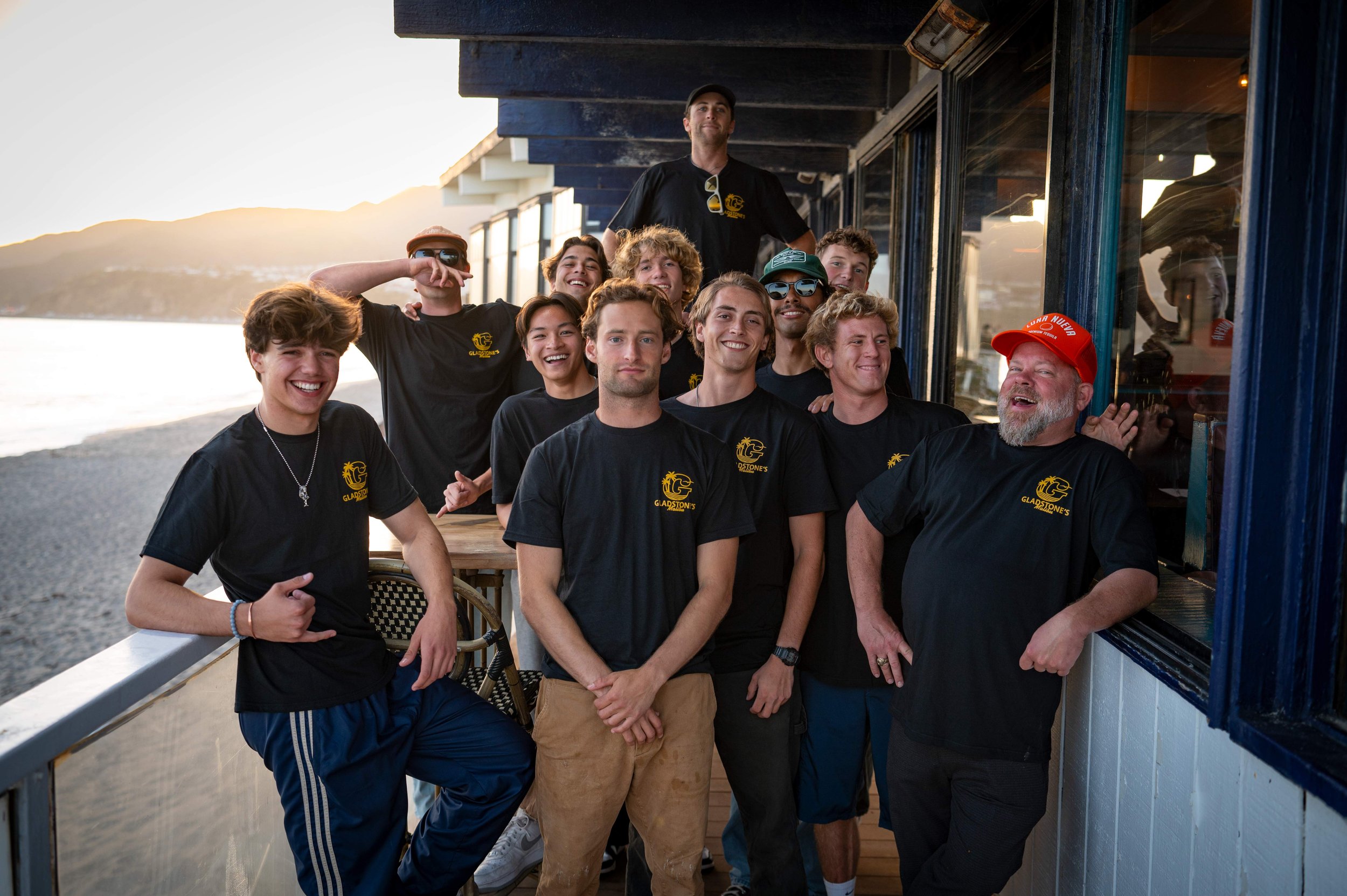 Group of young men and an older man smiling and posing on a deck during sunset, with mountains and water in the background, wearing matching black t-shirts and casual clothing.
