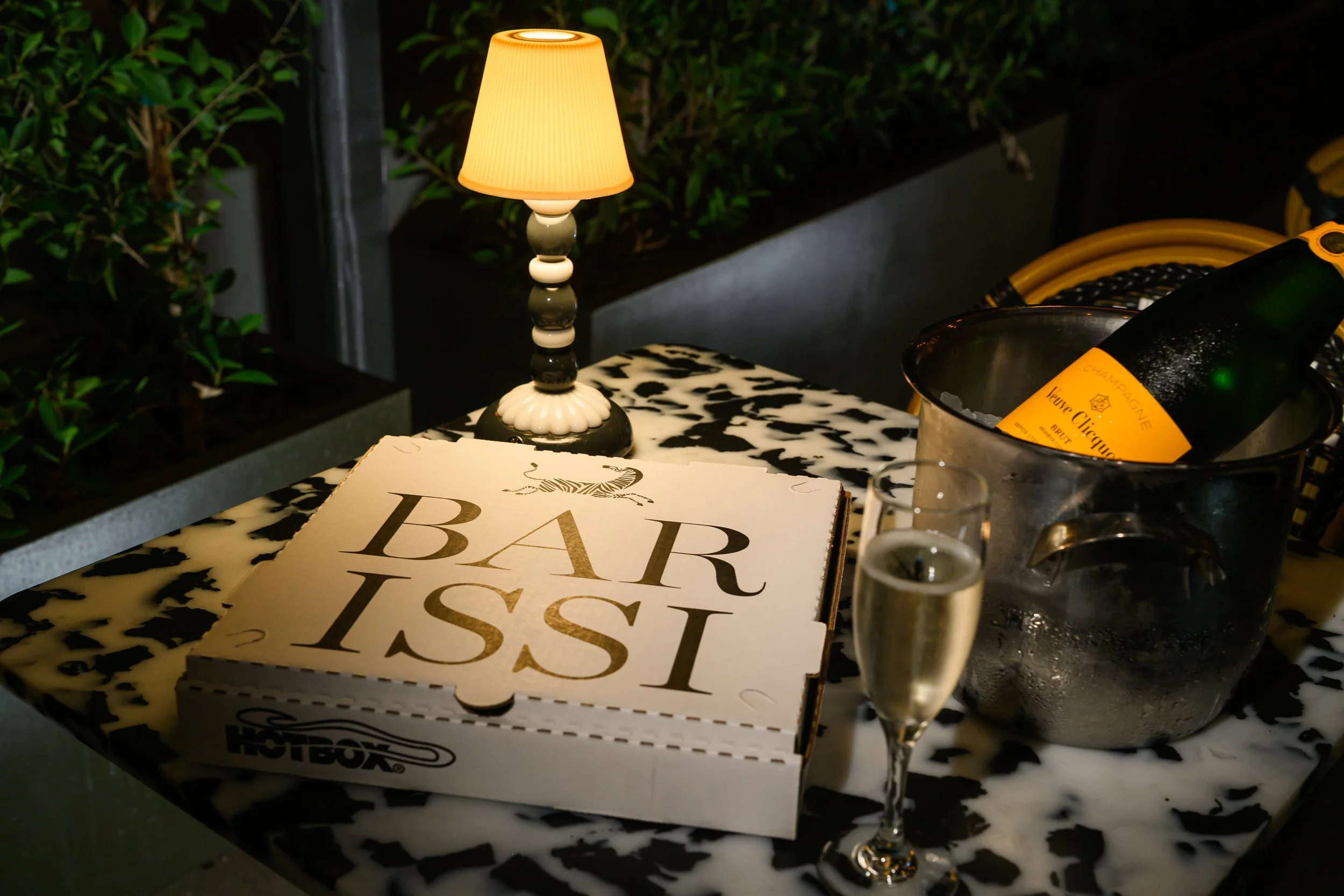 A small table with a black and white patterned surface holding a lit beige lampshade table lamp, a pizza box labeled "BAD FISH," a bottle of champagne in an ice bucket, and a glass of sparkling water. There are plants and dim lighting in the backgrou