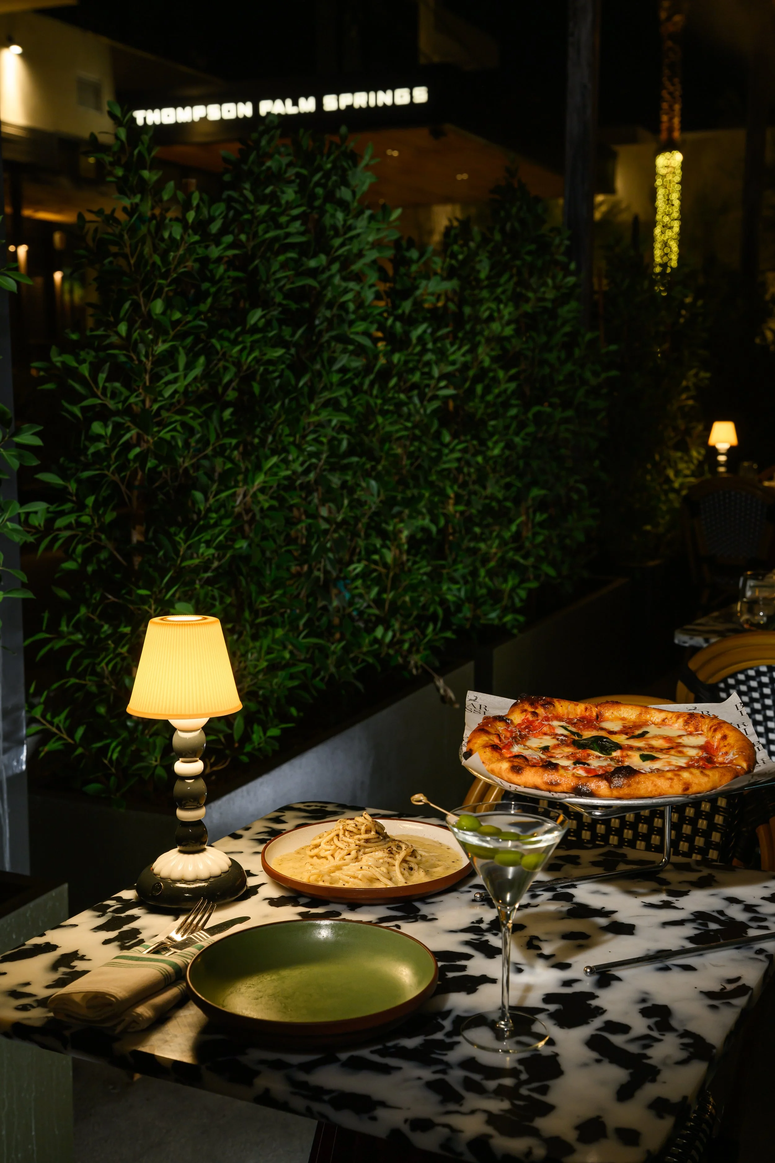 A table set with pasta, a pizza on a stand, a martini glass with olives, and a lamp, with a green leafy backdrop and ambient lighting.