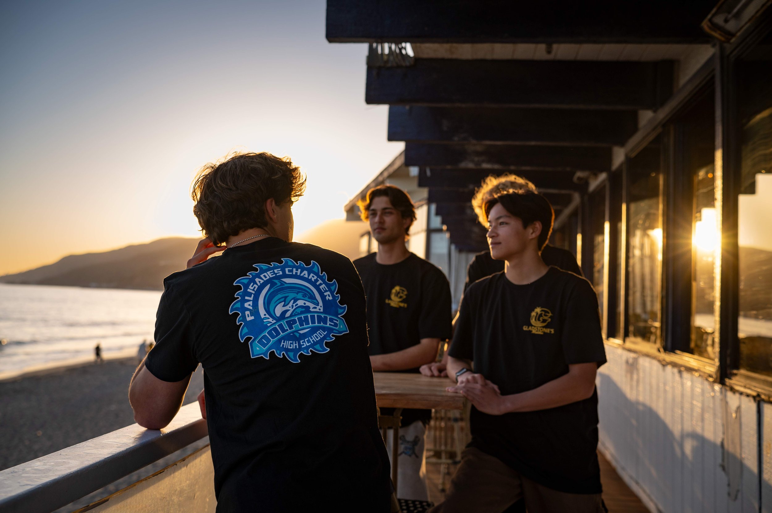 Four young men are standing on a wooden deck near the waterfront during sunset, engaged in conversation, with mountains and the ocean in the background.