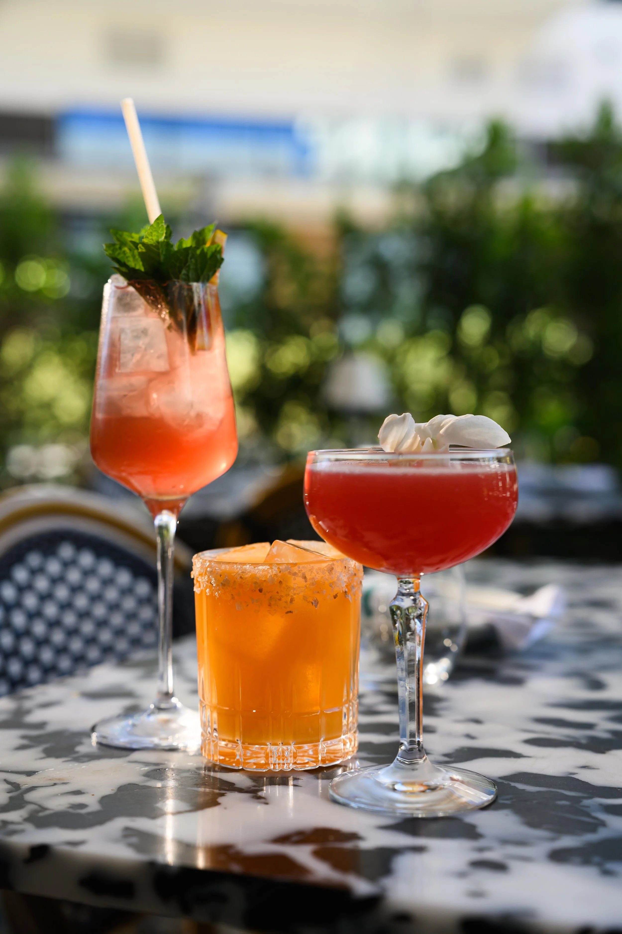 Collection of colorful cocktails on a table outside, with blurred greenery and buildings in the background.