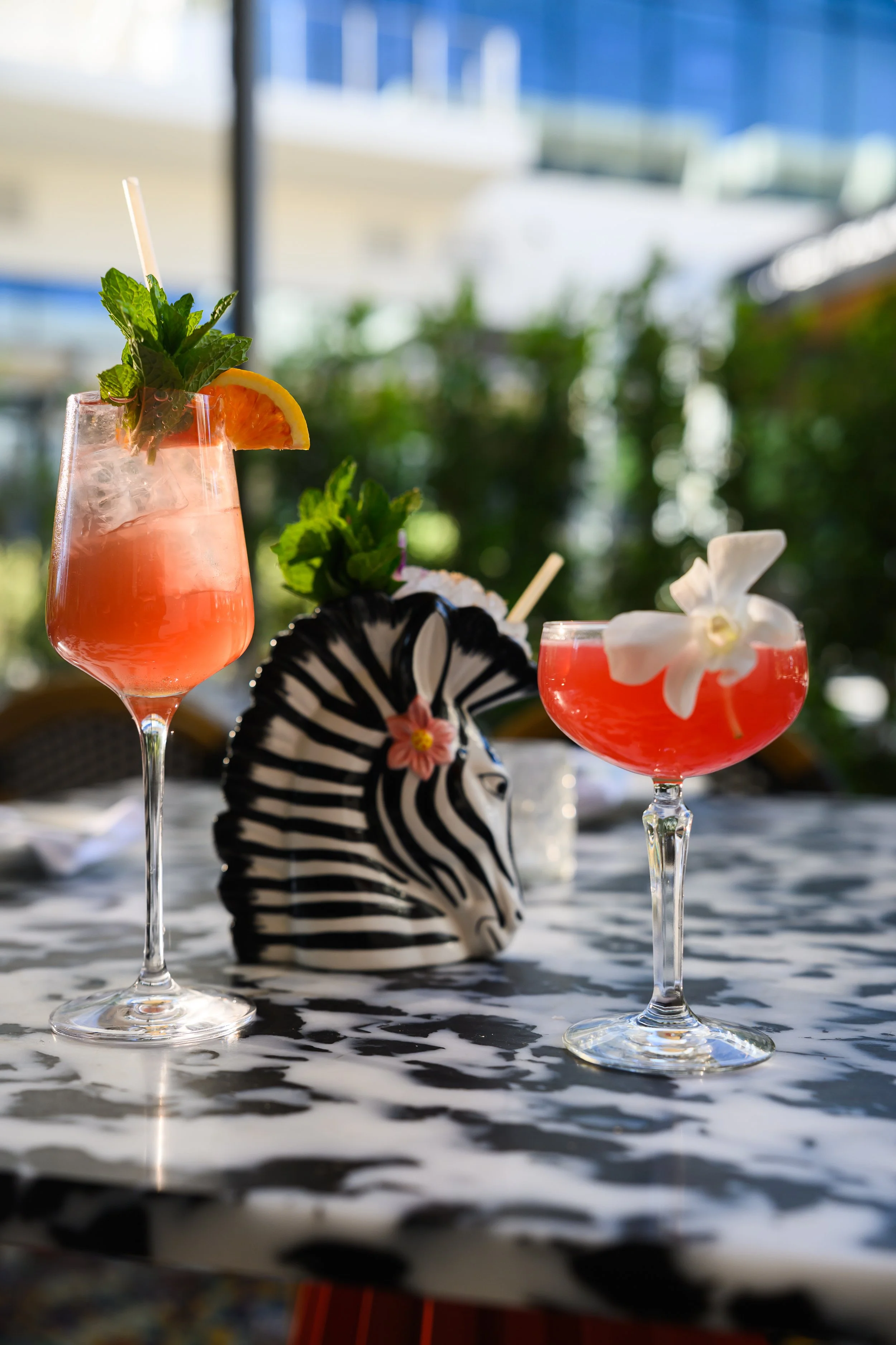 Two pink cocktails in elegant stemmed glasses with garnishes of mint, orange slices, and edible flowers on a patterned table surface, with a black-and-white zebra-striped decorative object and greenery in the background, under a see-through umbrella.