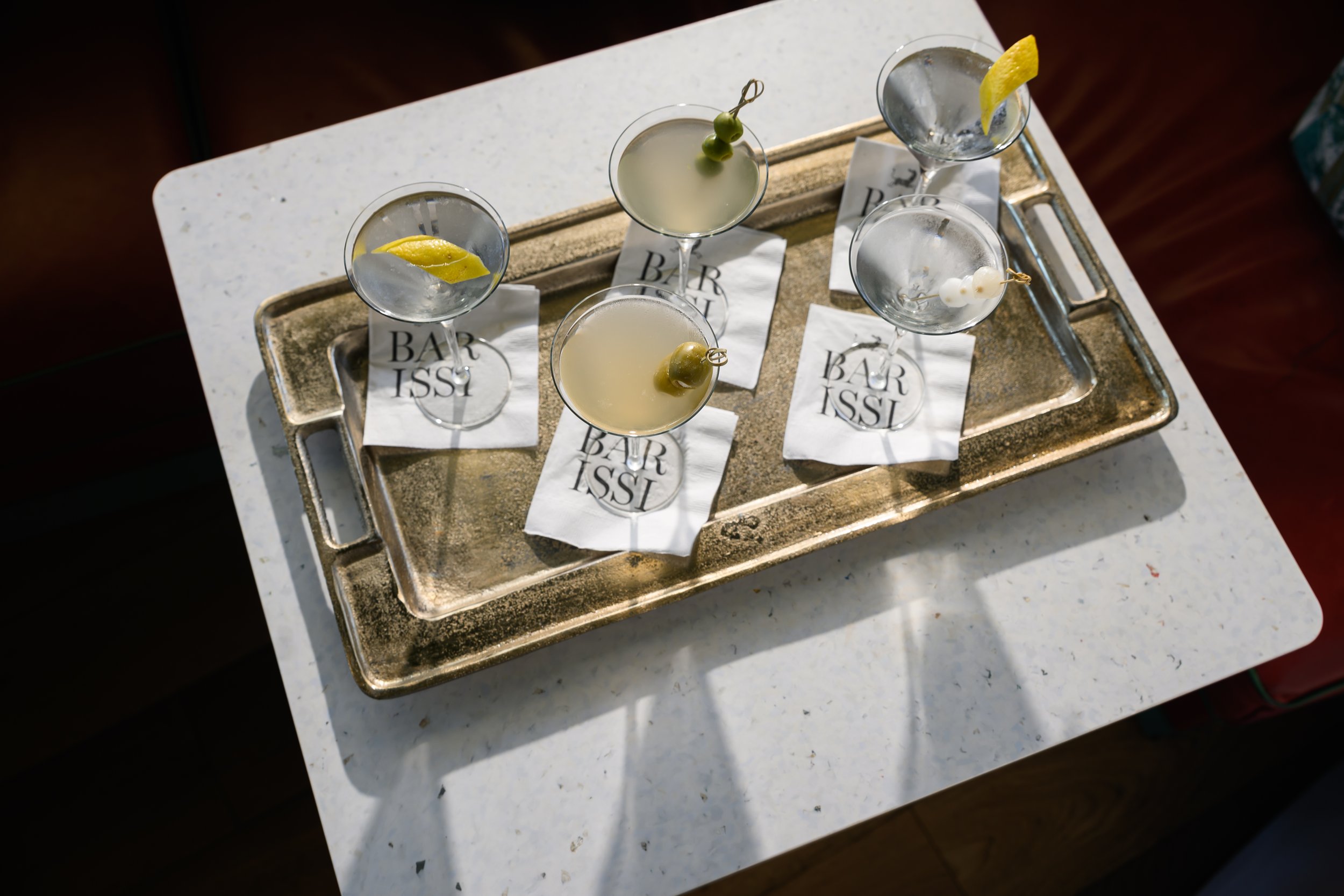 A silver tray with five cocktail glasses containing various garnished drinks, placed on a white table with Bar Issi napkins underneath.