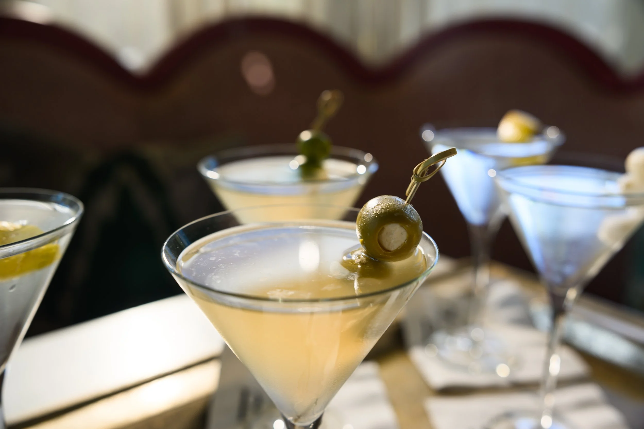 Multiple martini glasses filled with creamy white cocktails, each garnished with a green olive on a toothpick, set on a table with blurred background.