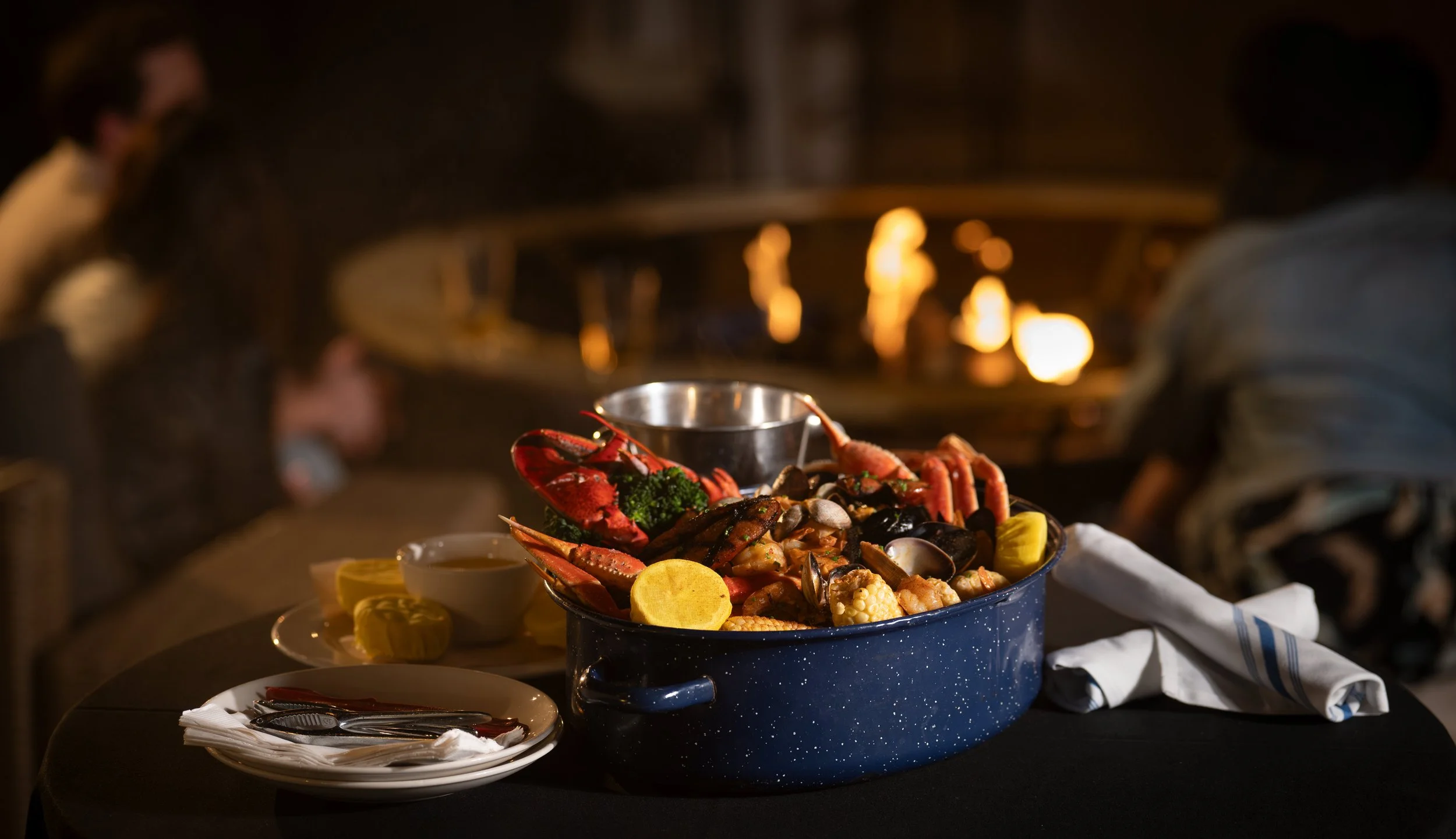 A seafood feast including lobster, shellfish, and vegetables served in a large blue pot, with lemons, butter, and utensils around it, set on a table in a dimly lit restaurant.