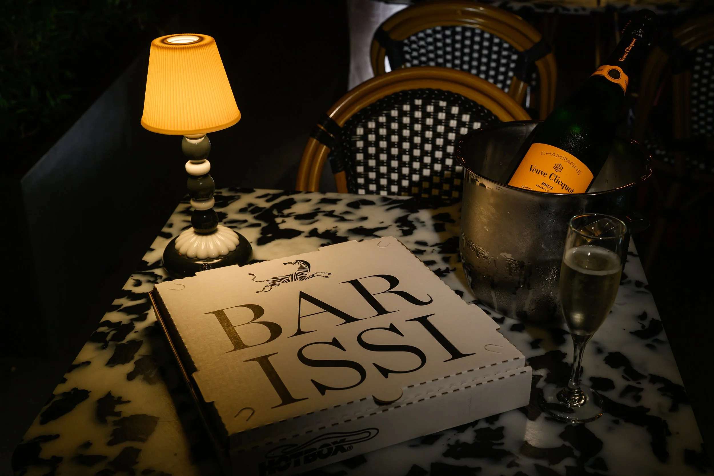 A table with a small lamp, a stack of books titled 'Bar', a cheese platter, two glasses of champagne, and a bottle of Veuve Clicquot champagne in a silver ice bucket. There are chairs with black and white checkered seats and rattan backs in the background.