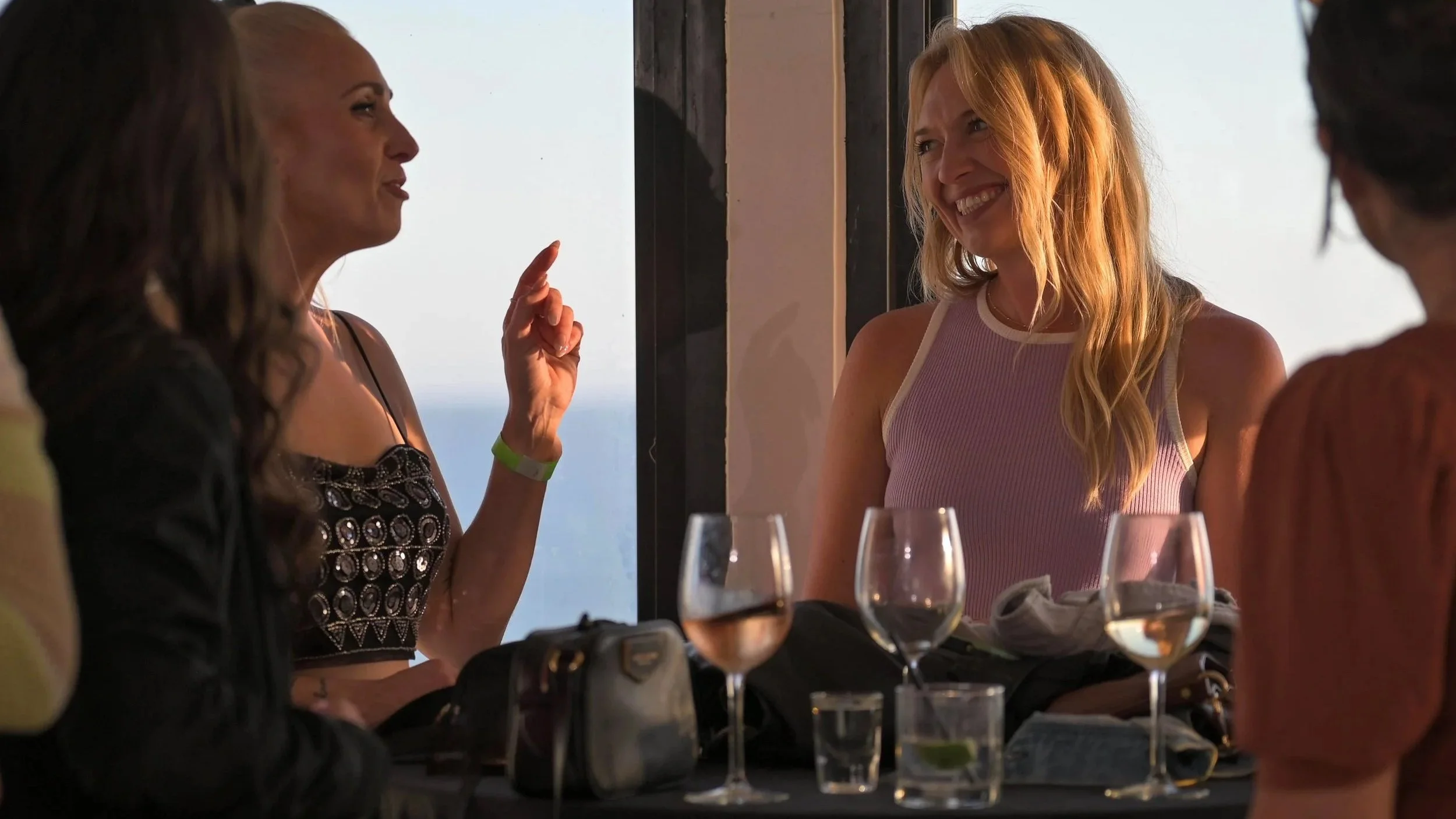 People socializing at a restaurant with glasses of wine, during daytime, with a view of the sky through large windows.