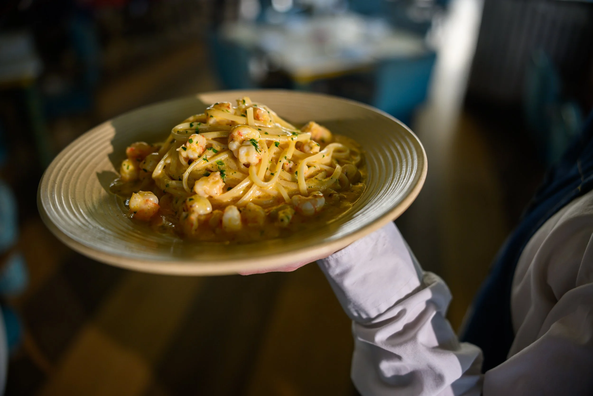 Person in white shirt holding a plate of shrimp pasta with parsley, in a dimly lit restaurant or kitchen setting.