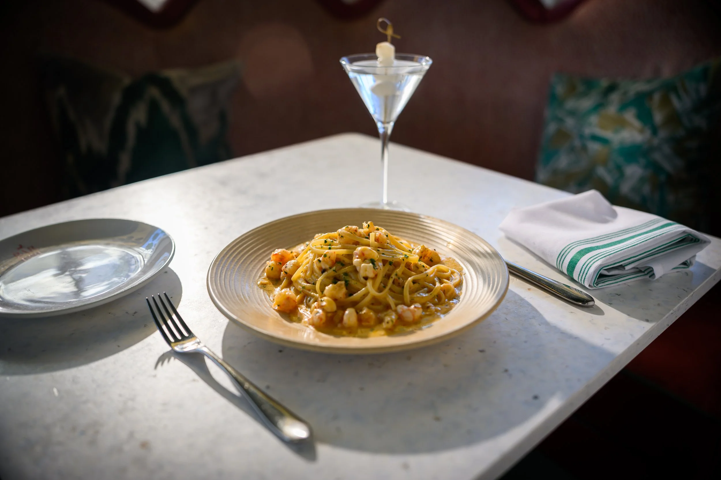 A plate of shrimp pasta with a creamy sauce on a white marble table, with a fork, a glass of water, a plate, and a napkin set on the table.