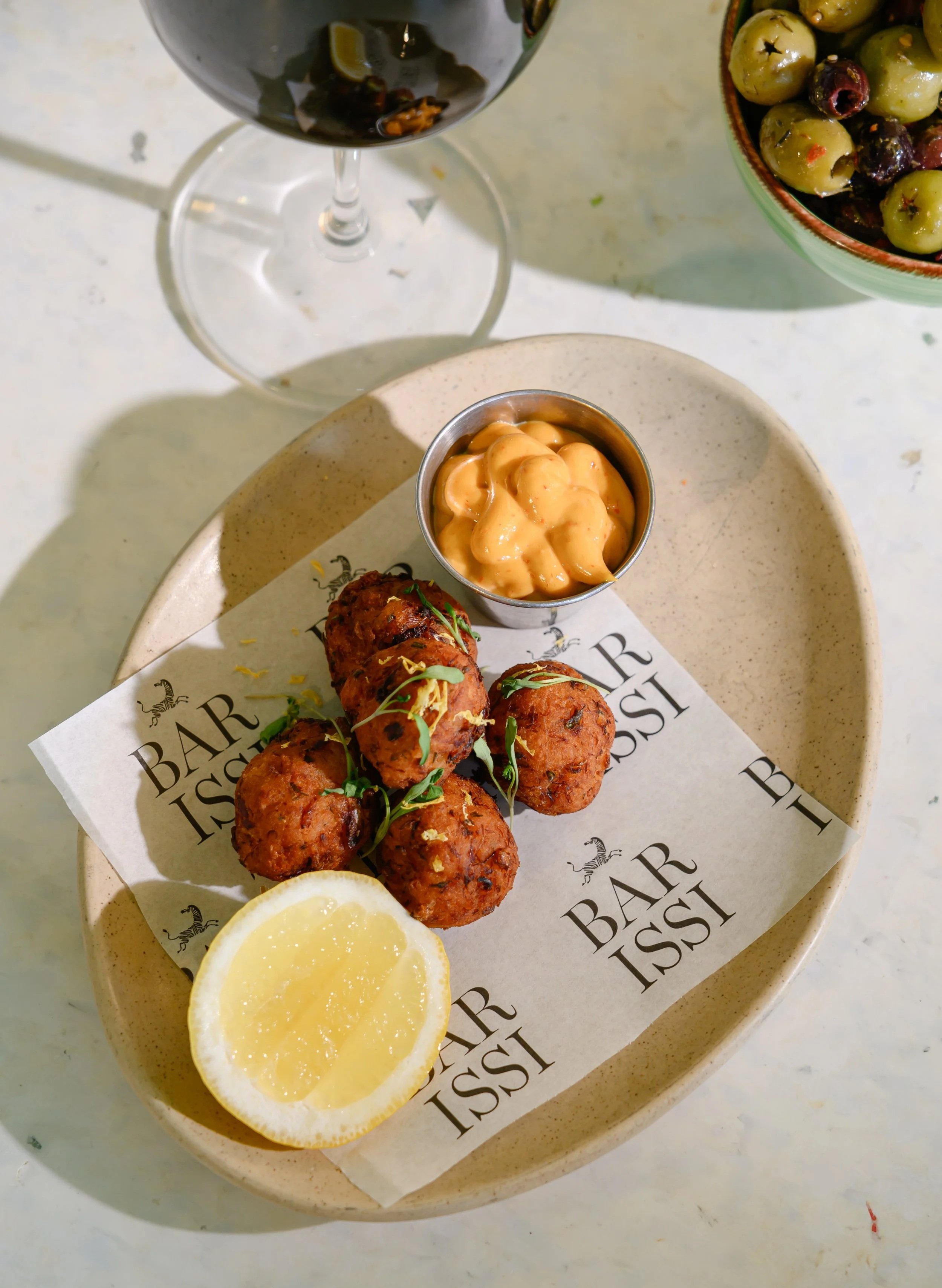 A plate with four fried crab cakes garnished with microgreens, two lemon slices, a small metal cup of creamy sauce, and a piece of lemon on the side. In the background, there is a bowl of green olives with red pepper flakes and a glass of dark red wine with a lemon wedge. The plate is on a white surface with BARE ISS insignia on a paper liner.