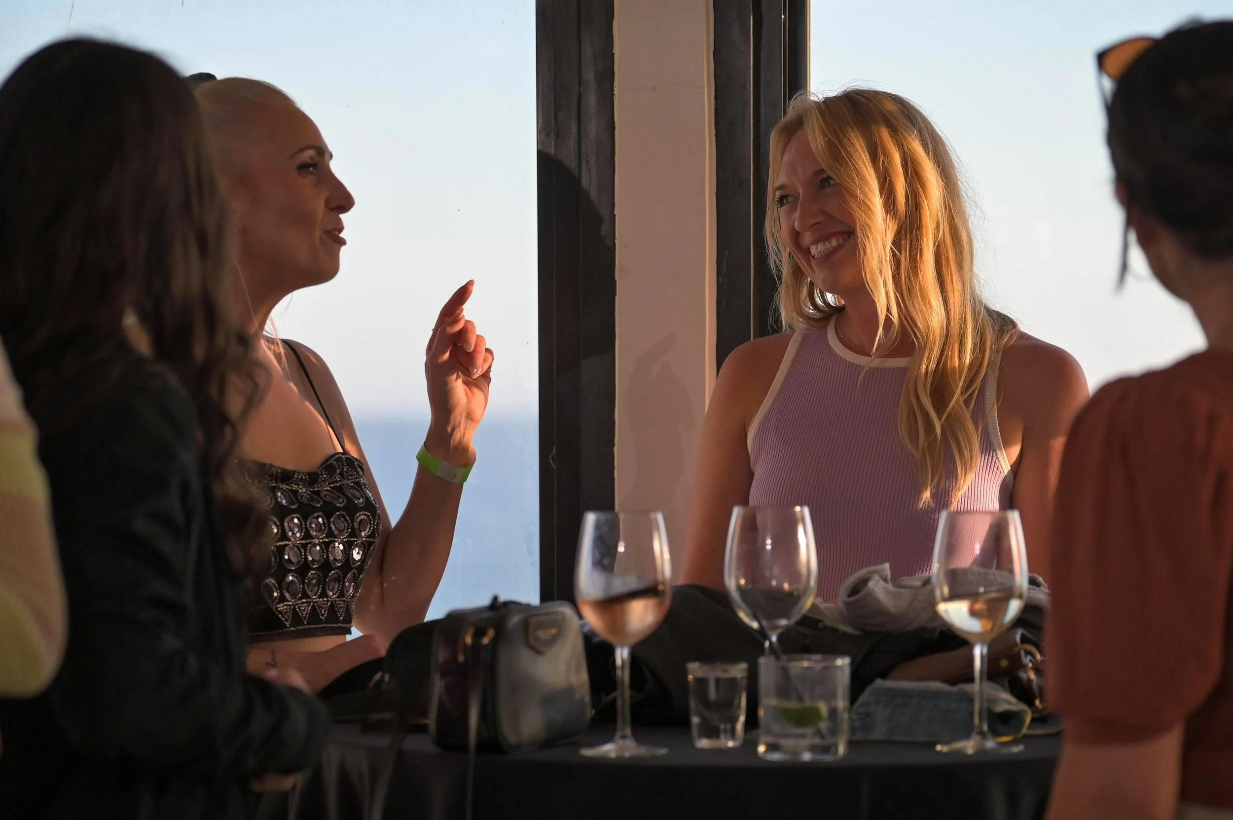 Four women are sitting at a table with glasses of wine, engaged in conversation. One woman is smiling, and another woman appears to be speaking, with her finger raised. The setting looks like a restaurant or a social gathering with a window showing a