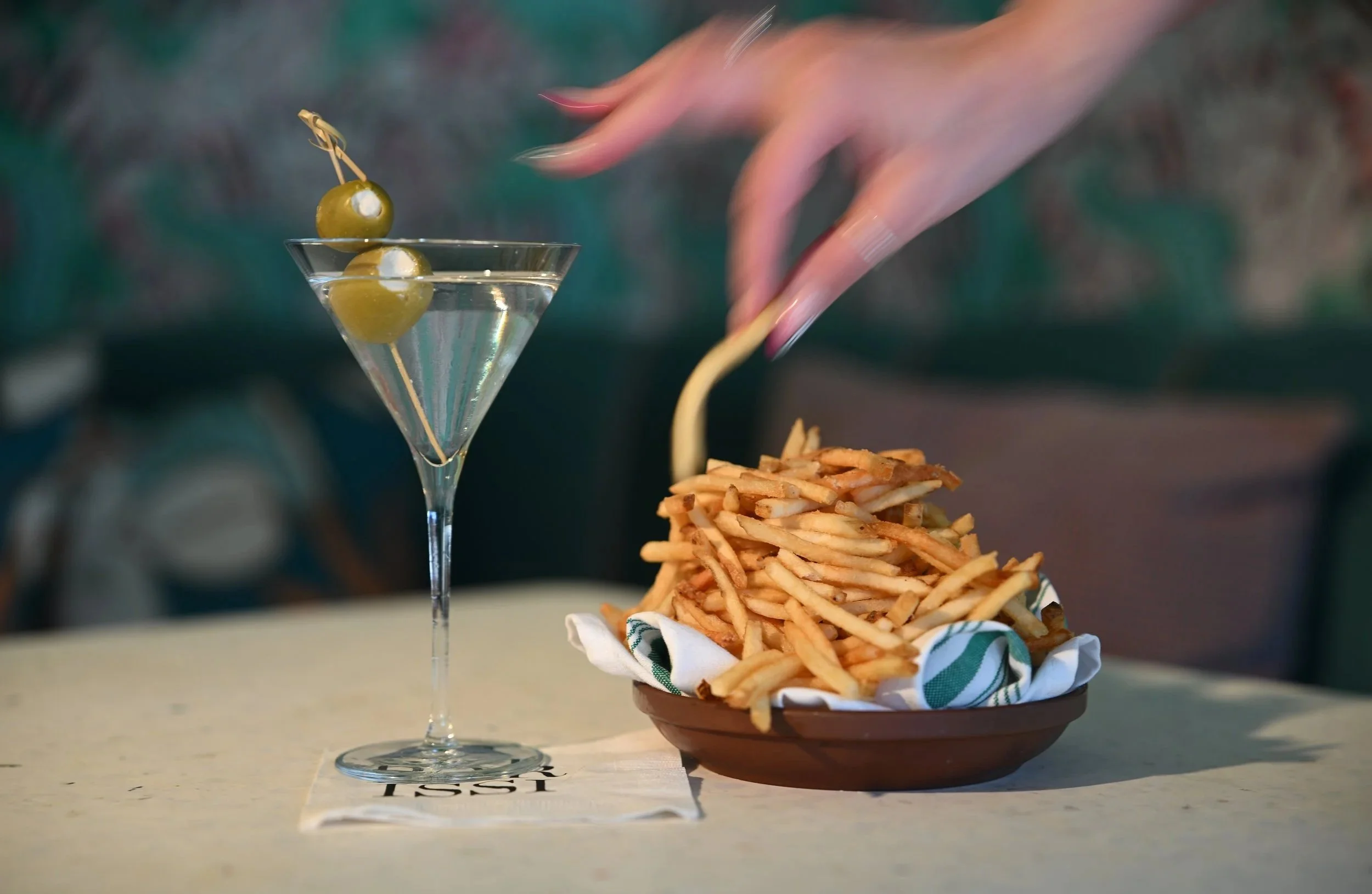 A martini glass with green olives on a pick is placed on a table next to a basket of crispy French fries with a cloth napkin. In the background, a person's arm is reaching for the fries.