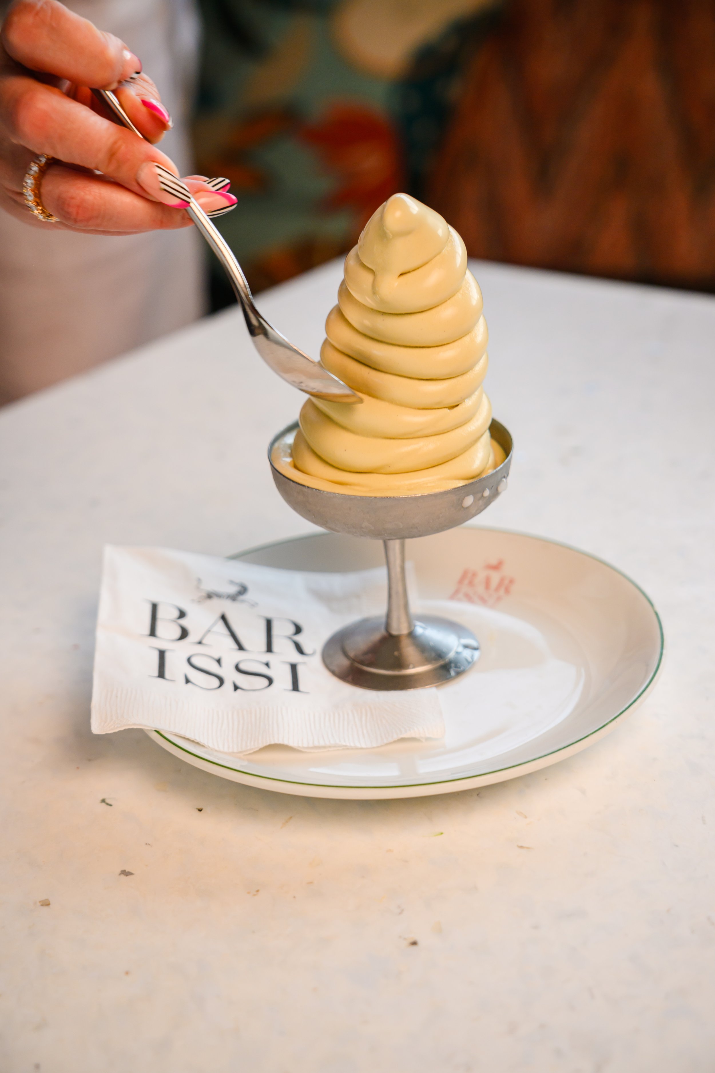 A soft serve ice cream cone in a silver cup on a white plate, with a napkin that reads 'BAR IST' underneath, on a white table.