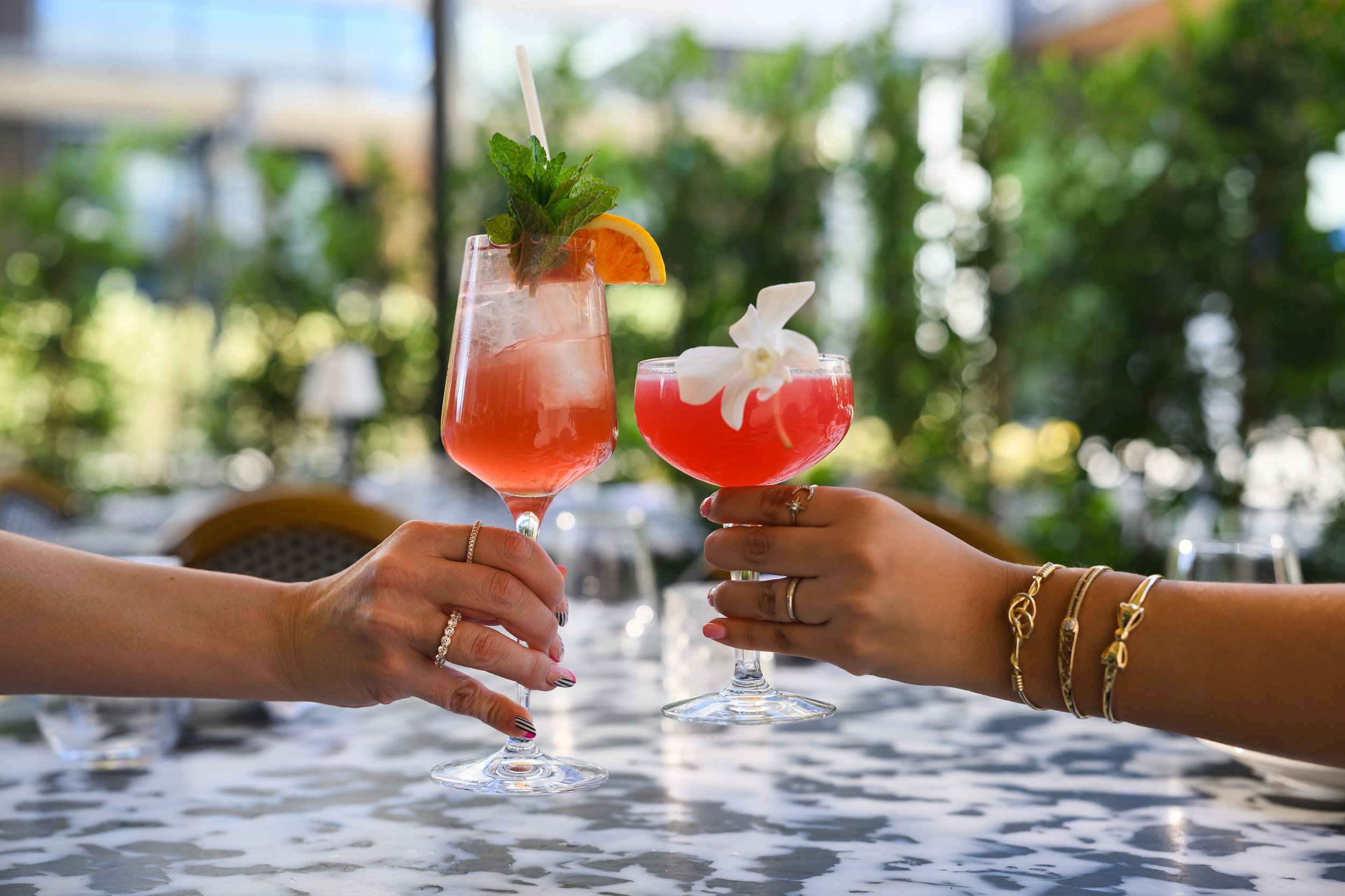 Two people are clinking glasses of pink cocktails with garnishes, including an orange slice, mint leaves, and a white flower, in a bright outdoor setting.