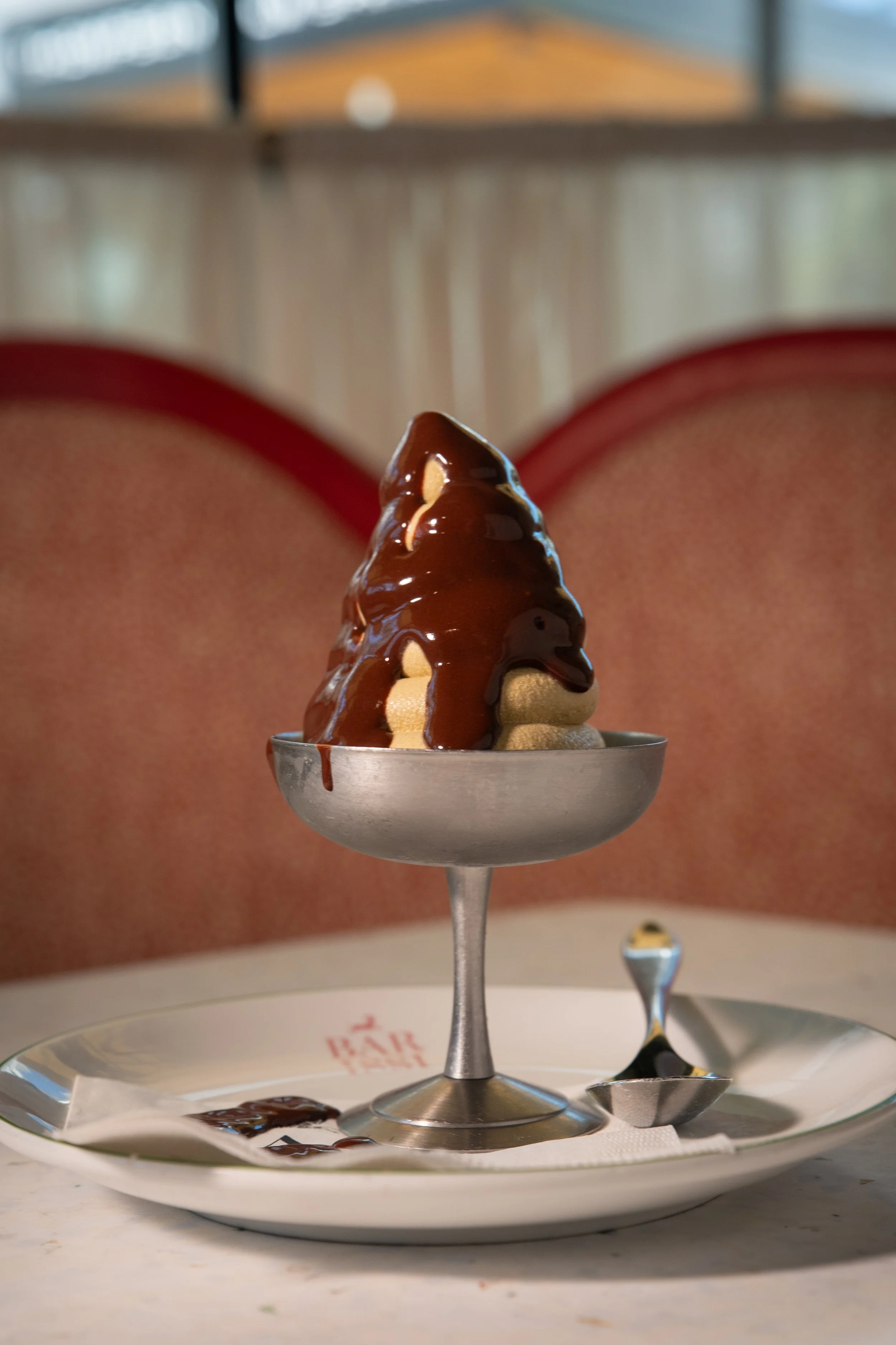 Dessert with chocolate syrup and nuts served on a metal stand, placed on a white plate with a spoon. Background shows a person wearing red shorts.