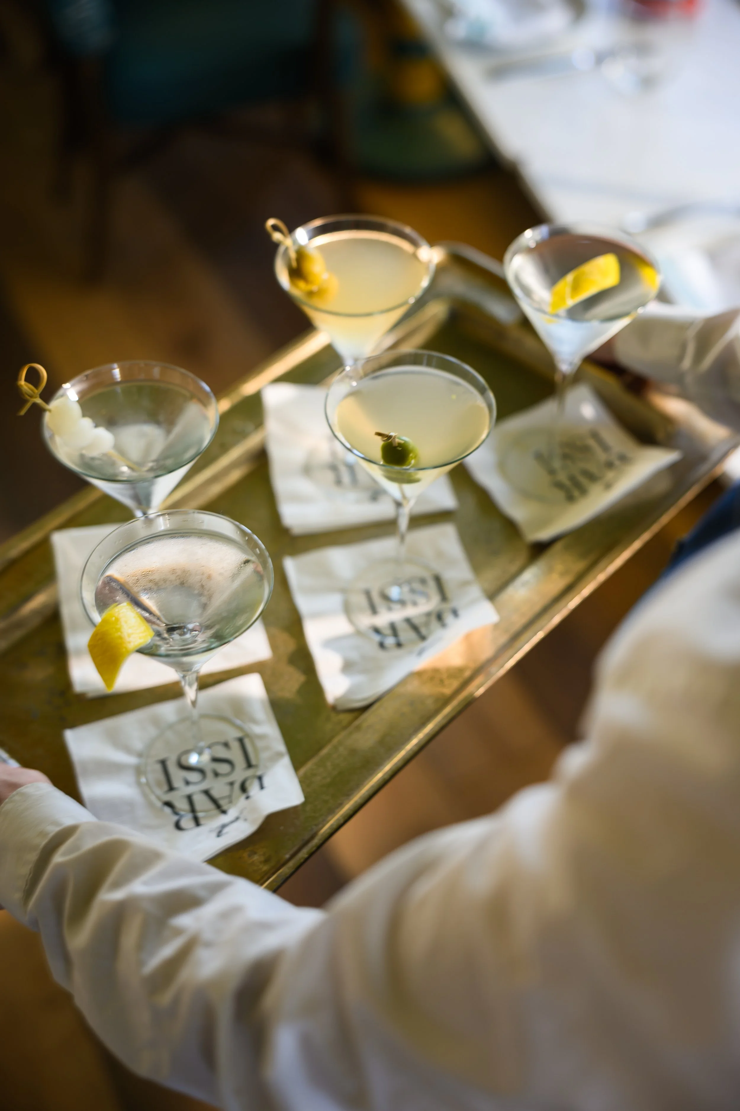 Multiple martini glasses with lemon twists and olives on white napkins, served on a wooden tray.