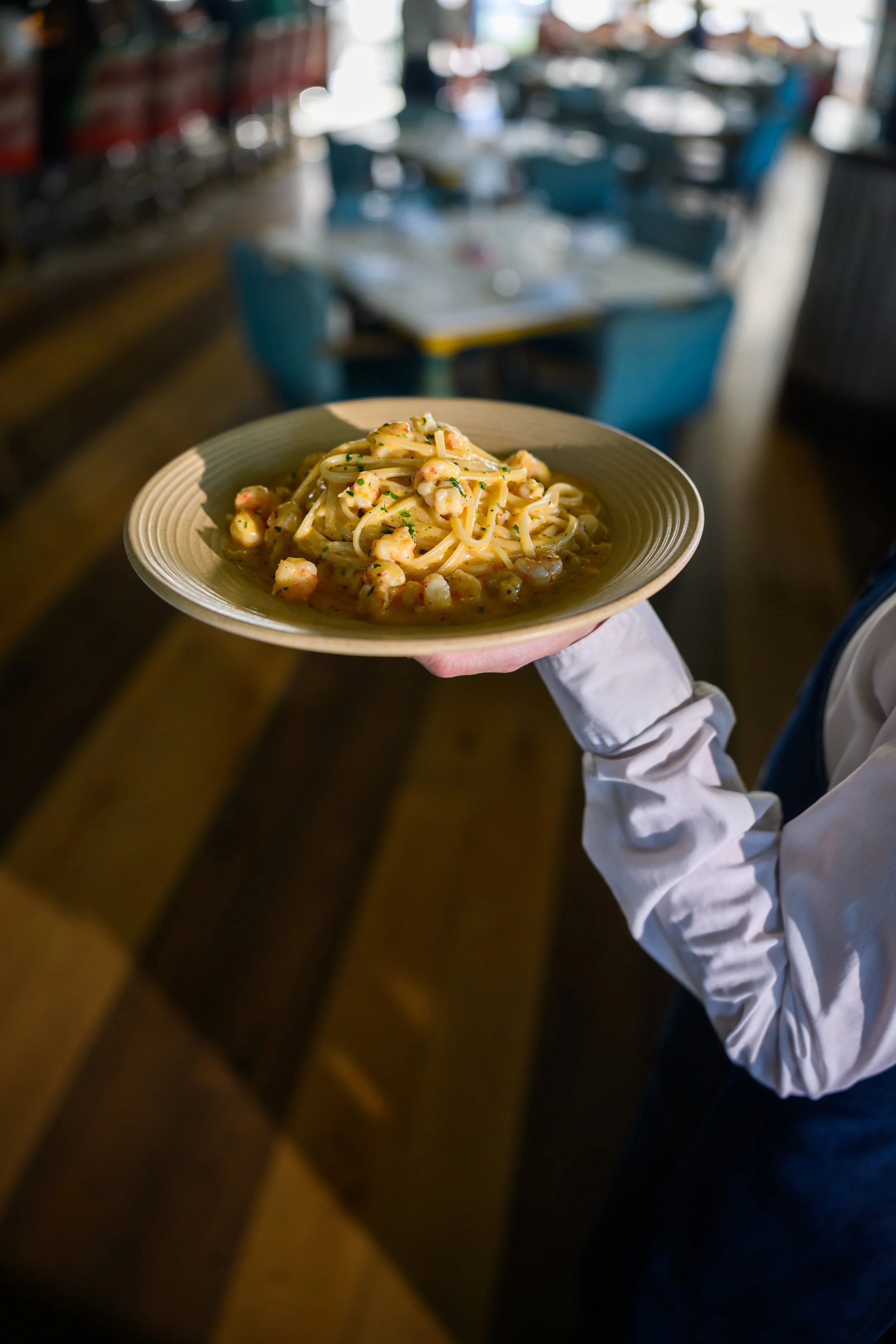 A person holding a plate of seafood pasta in a restaurant with blue chairs and tables in the background.