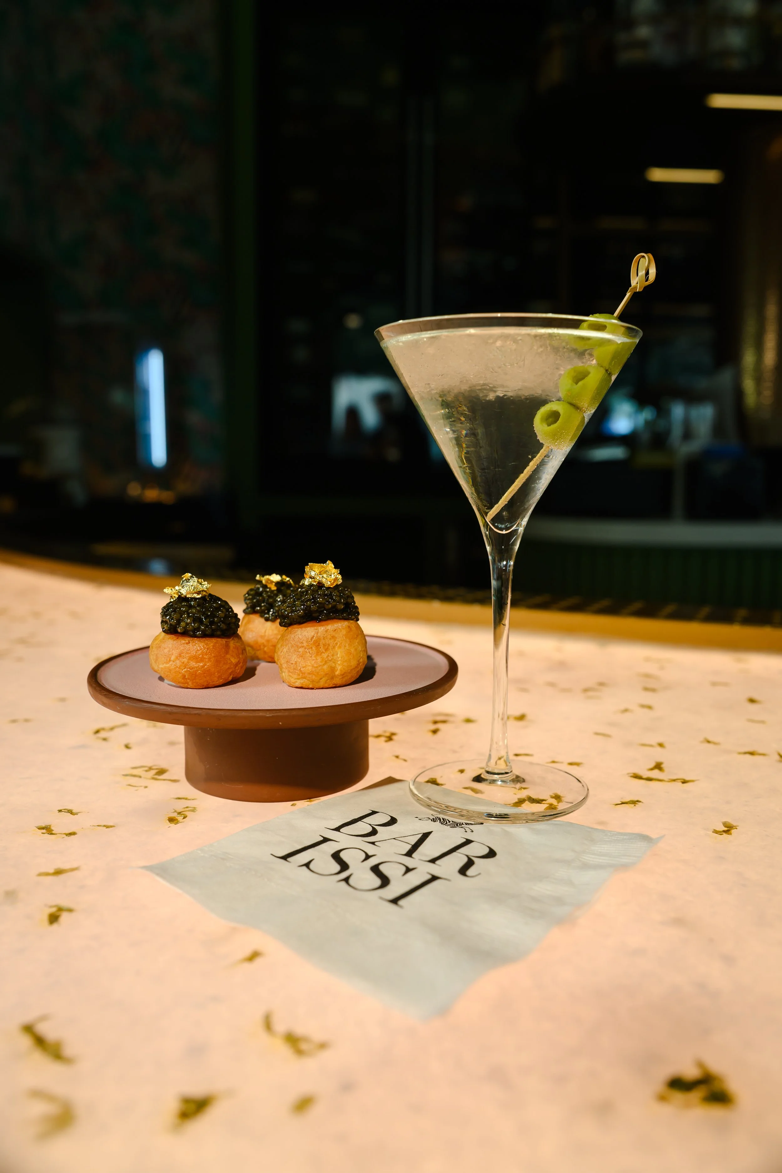A martini glass with green olives on a toothpick, and three bite-sized appetizers topped with black caviar and edible gold leaf, placed on a pink plate on a table with a paper that says 'Bar Book' at the bottom.