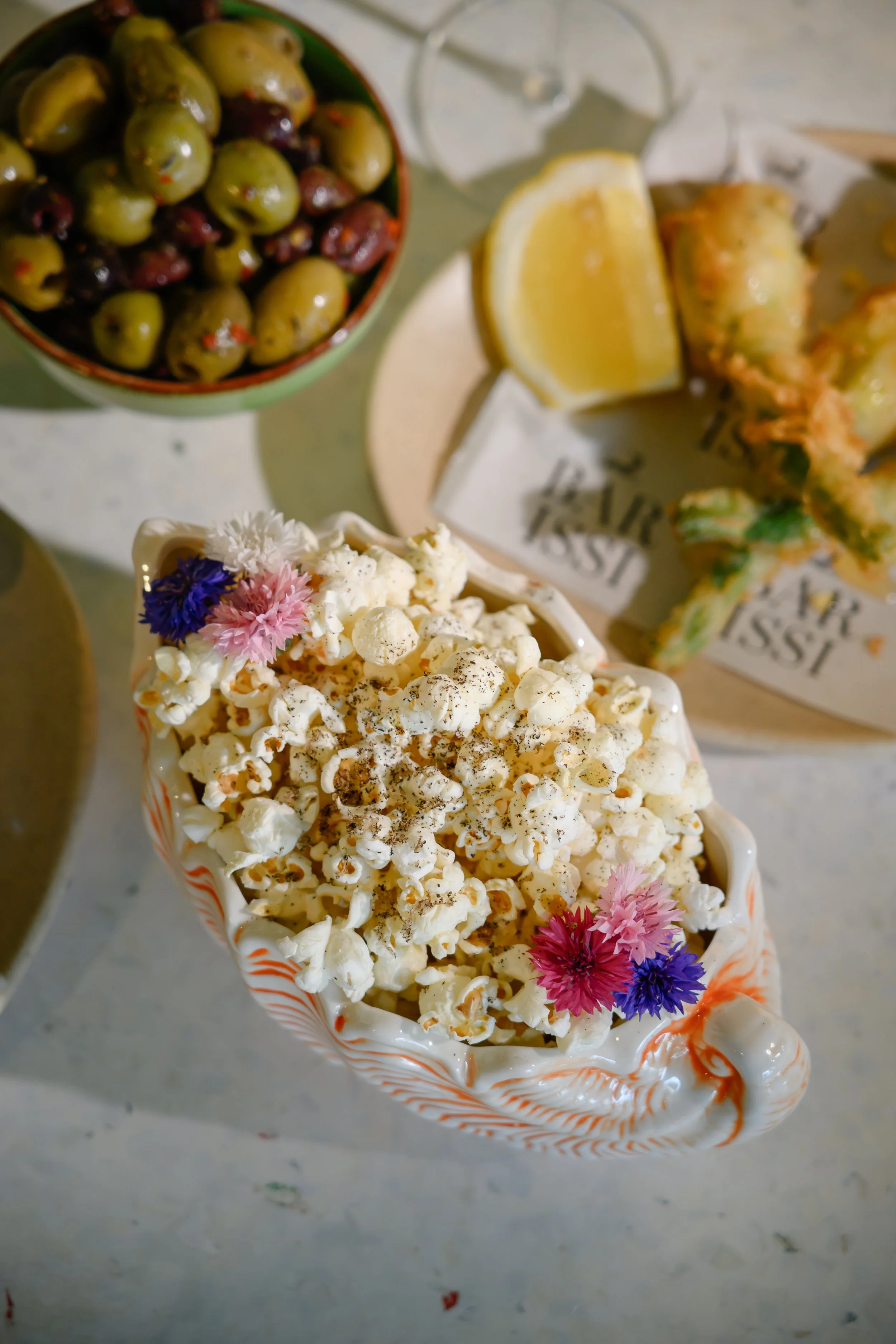 A bowl of popcorn topped with black pepper and decorated with pink and purple flowers. Behind it, a bowl of green and black olives and a lemon wedge are visible on a table.