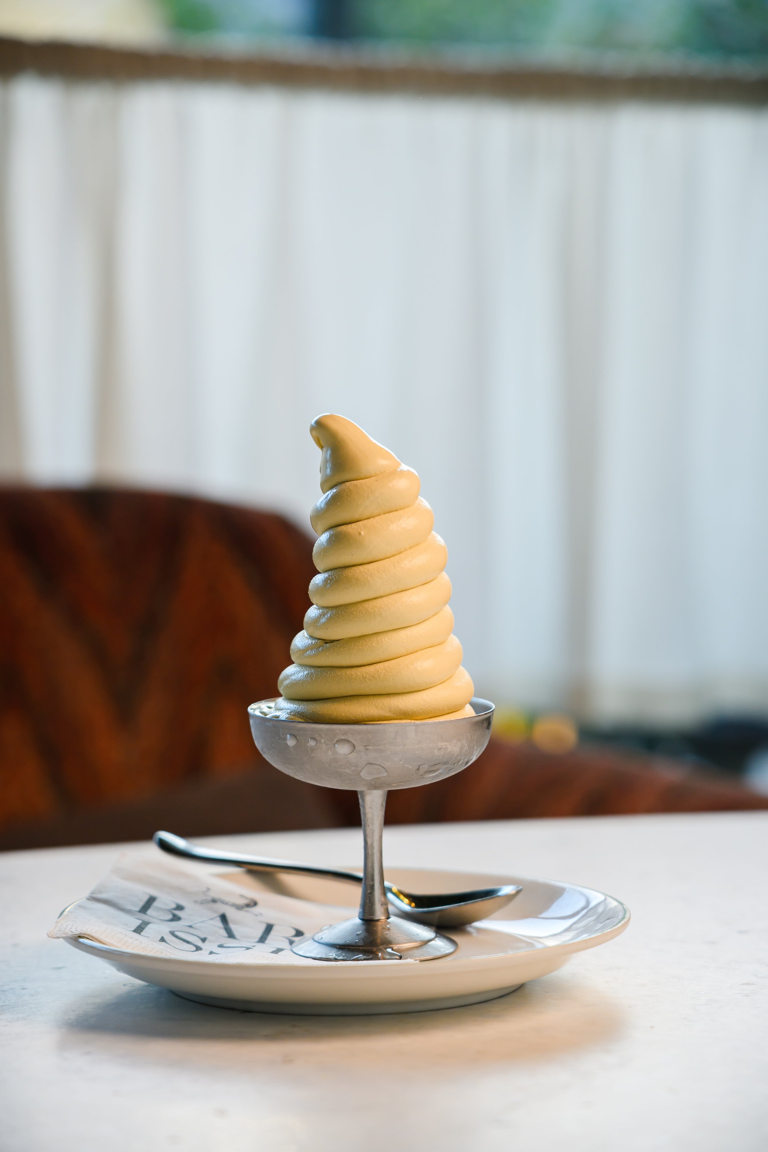 A tall glass dish of soft serve ice cream with a swirl shape and a yellowish color, placed on a white plate with a spoon. There's a paper napkin under the plate with partially visible text. In the background, out of focus, is a brownish fabric or obj