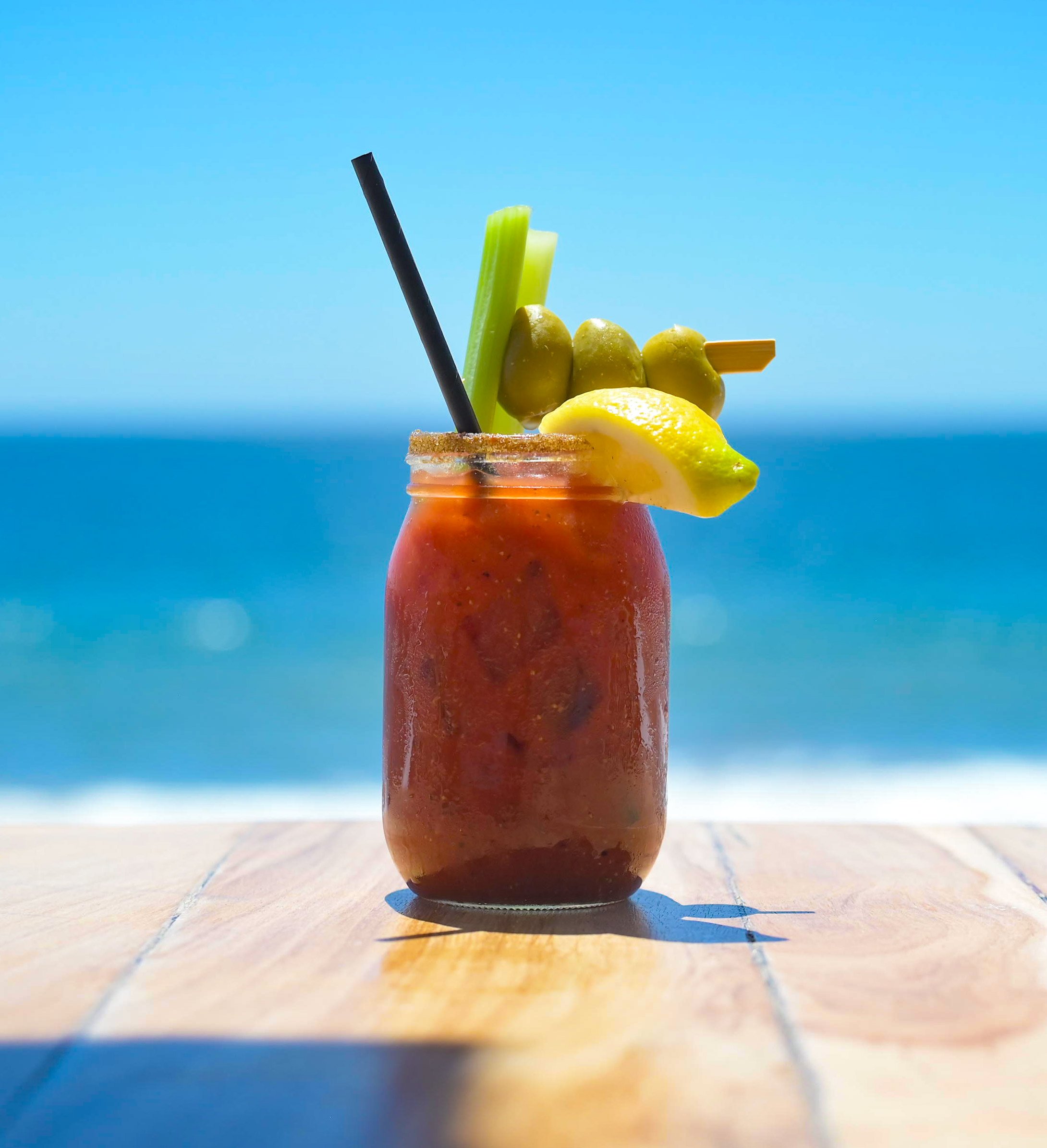 Close-up of a colorful cocktail with green olives, lemon wedges, celery, and a slice of lemon garnished on the rim of a glass, with a blue sky and ocean in the background, and a wooden surface below.