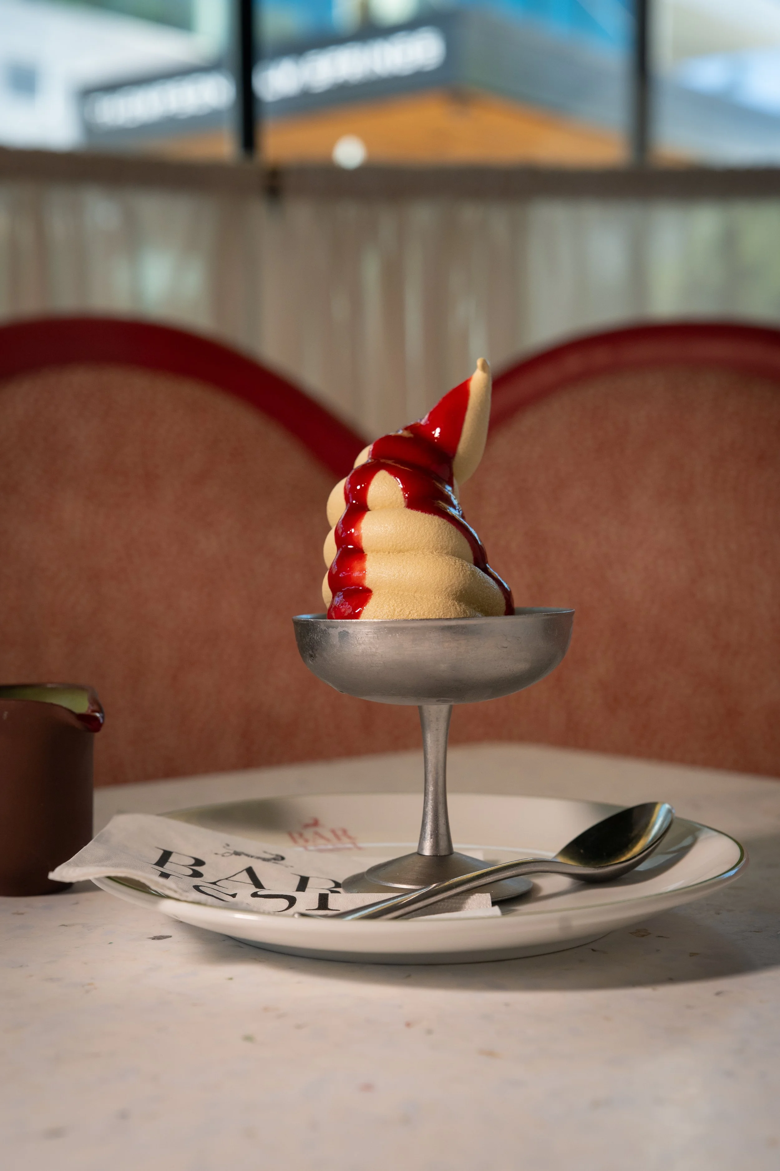 Frozen soft serve ice cream with red syrup drizzled on top, served in a silver metal cup on a white plate with a spoon. Behind it is a blurred background of a cafe or restaurant setting.