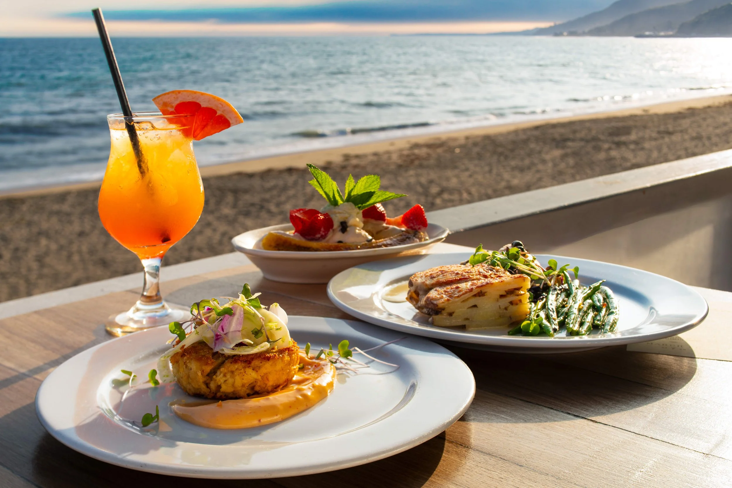 A seaside restaurant table with various plates of food including salads, pasta, and a grilled dish, along with a colorful cocktail with an orange slice and a straw, set against a backdrop of ocean waves and distant mountains at sunset.