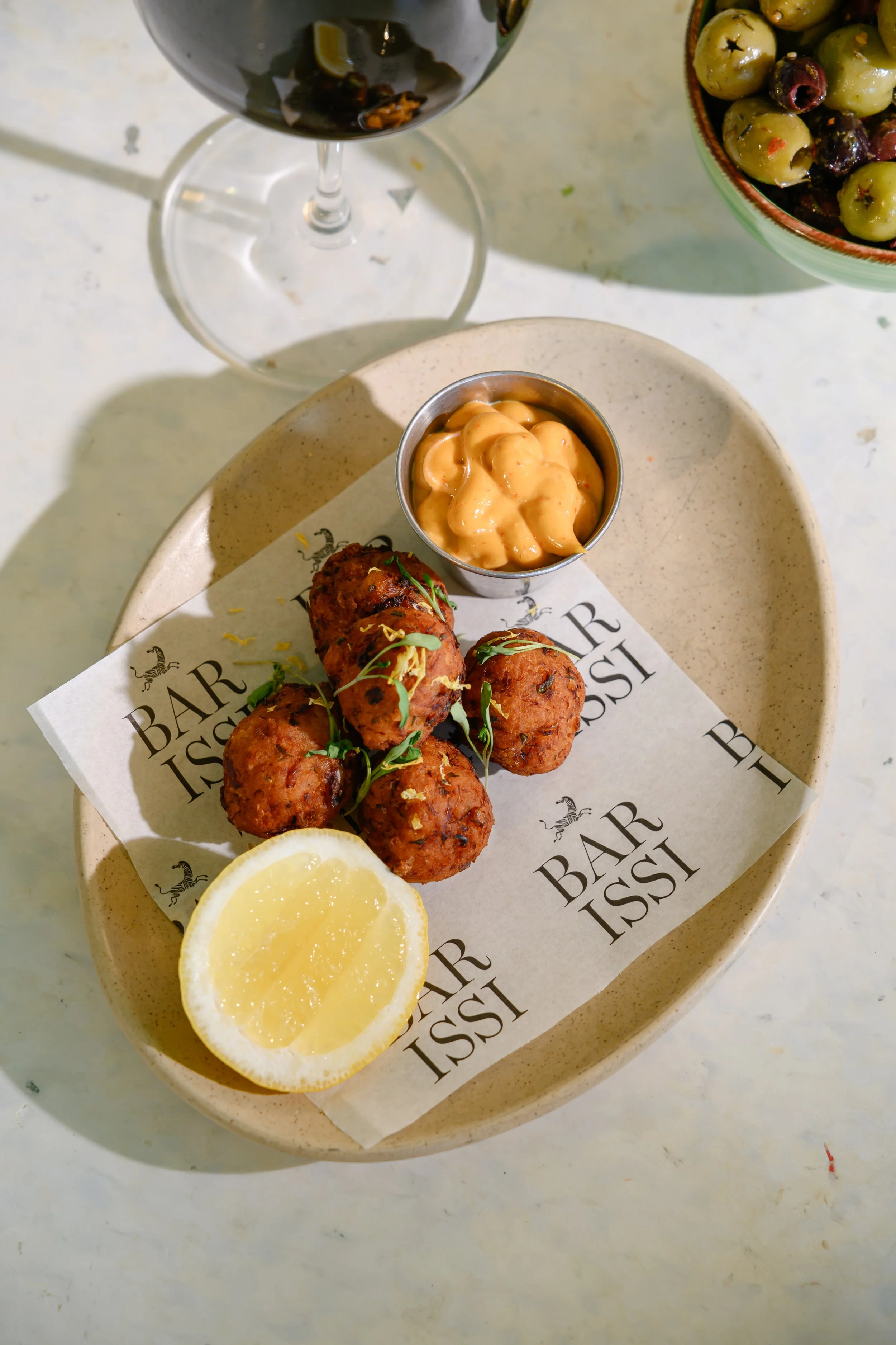 A plate with four falafel balls garnished with microgreens, a lemon wedge, and a small cup of creamy sauce. In the background, there is a bowl of green and black olives and a glass of red wine.