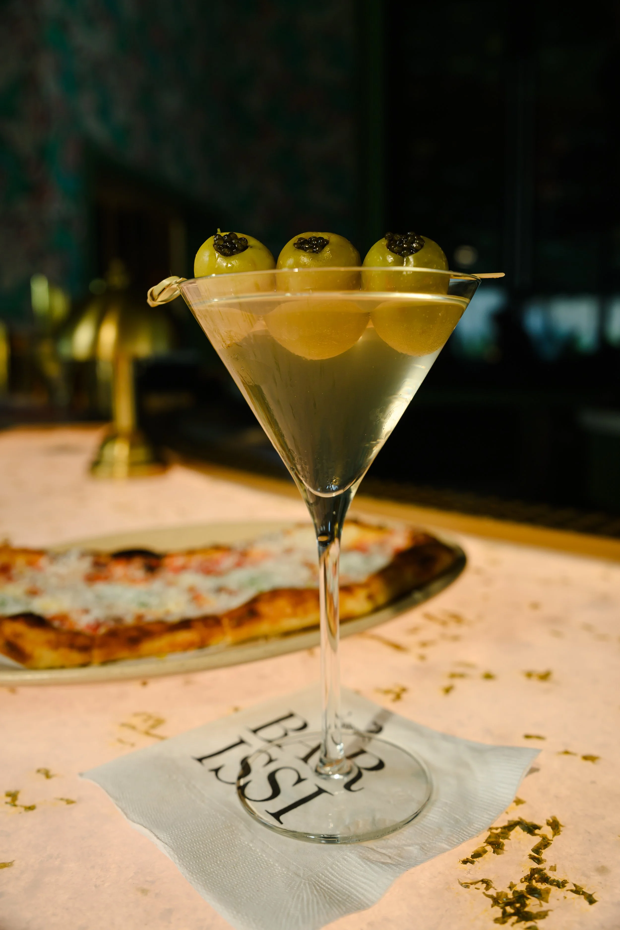A martini glass with three green olives garnished with black caviar. In the background, a pizza on a plate is slightly out of focus.