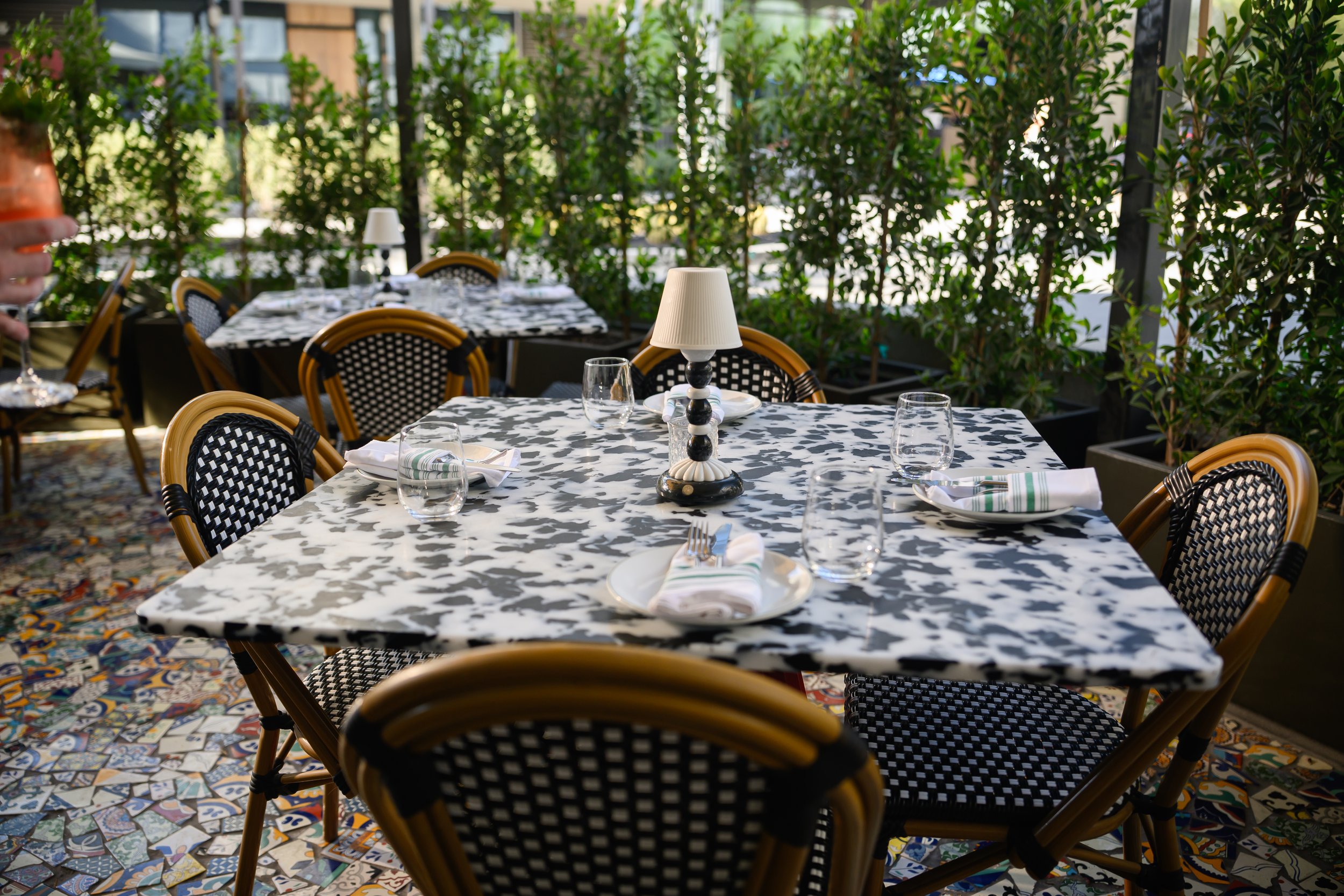 A restaurant table set with black and white patterned tablecloth, glassware, napkins, and a lamp in the center. Surrounding the table are black and tan woven chairs. There is decorative greenery and windows in the background, indicating an outdoor or