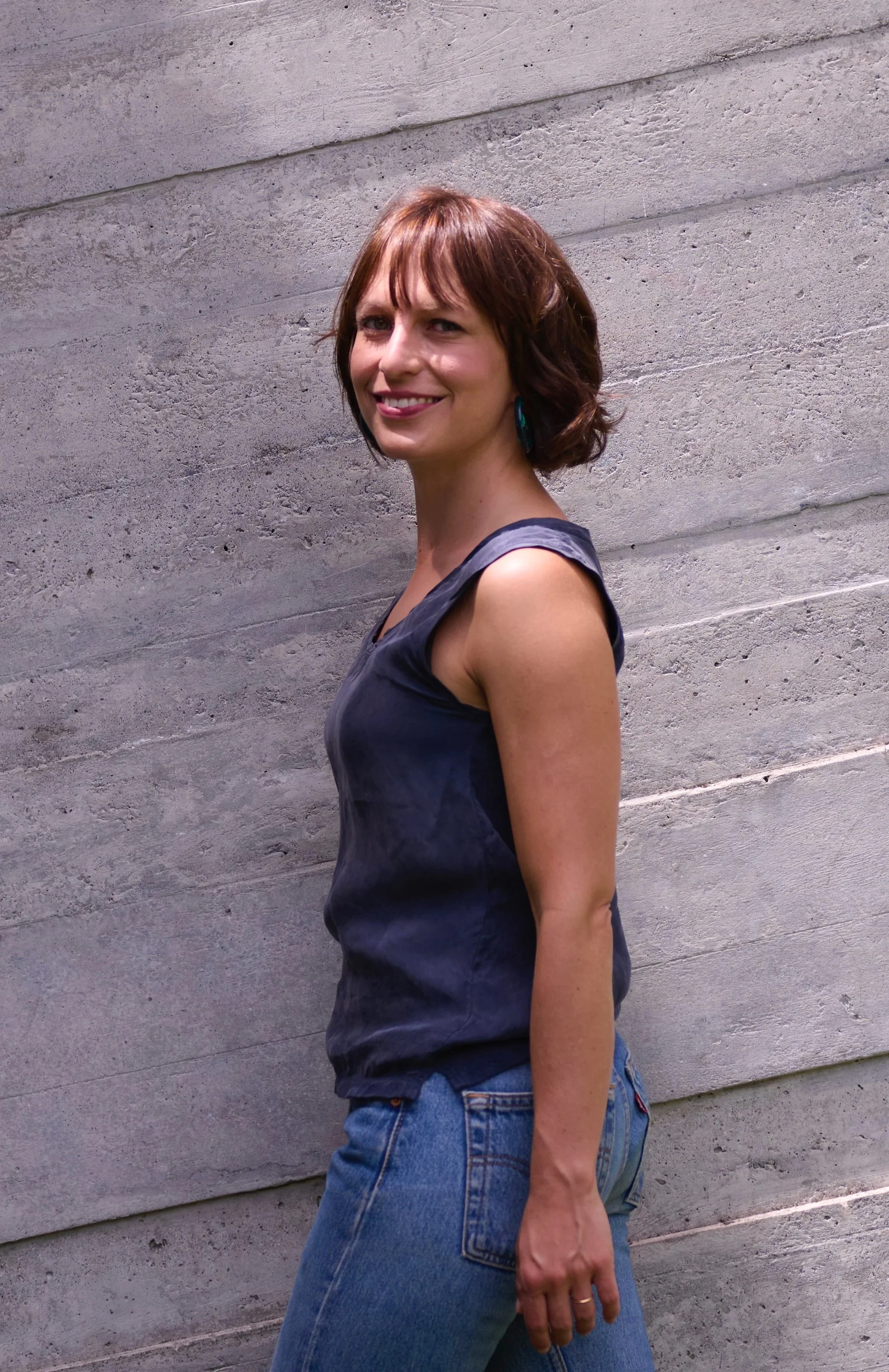 Roberta Diamond with short brown hair, wearing a black sleeveless top and blue jeans, standing sideways in front of a concrete wall, smiling at the camera.