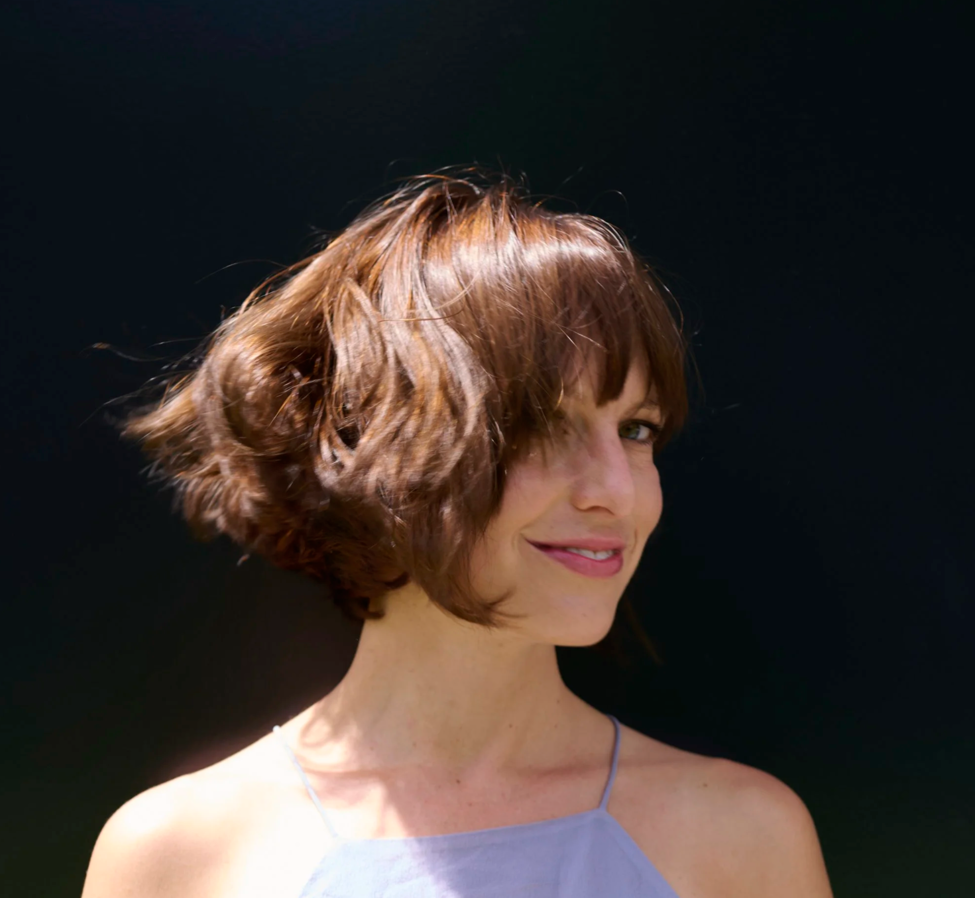 Close-up of a Roberta Diamond with short, wavy auburn hair wearing a light purple dress and a thin necklace, against a dark background.