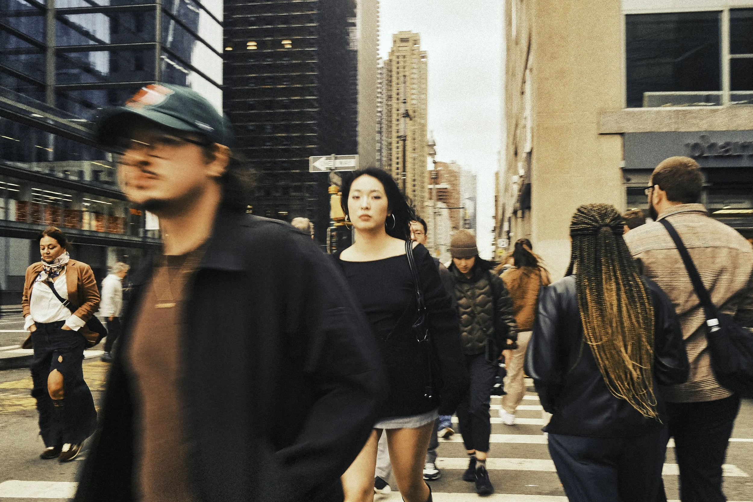 A crowded city street with people walking across a crosswalk. The scene is urban with tall buildings and skyscrapers, and the people include diverse individuals, some in casual clothing and others with accessories like hats and bags.