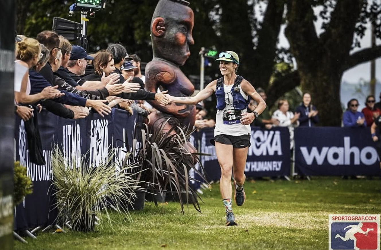 A female runner wearing a white shirt, black shorts, and a cap smiles and reaches out to shake hands with spectators along a race course. The crowd is clapping and reaching out to her. There is a large, colorful tribal-style sculpture in the backgrou