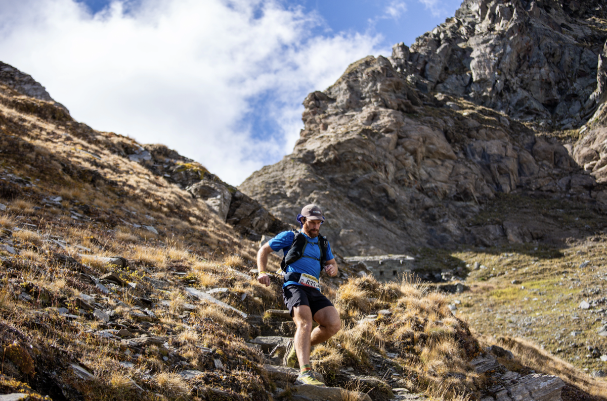 A man running down a rocky trail on a mountain with dry grass, under a partly cloudy sky.