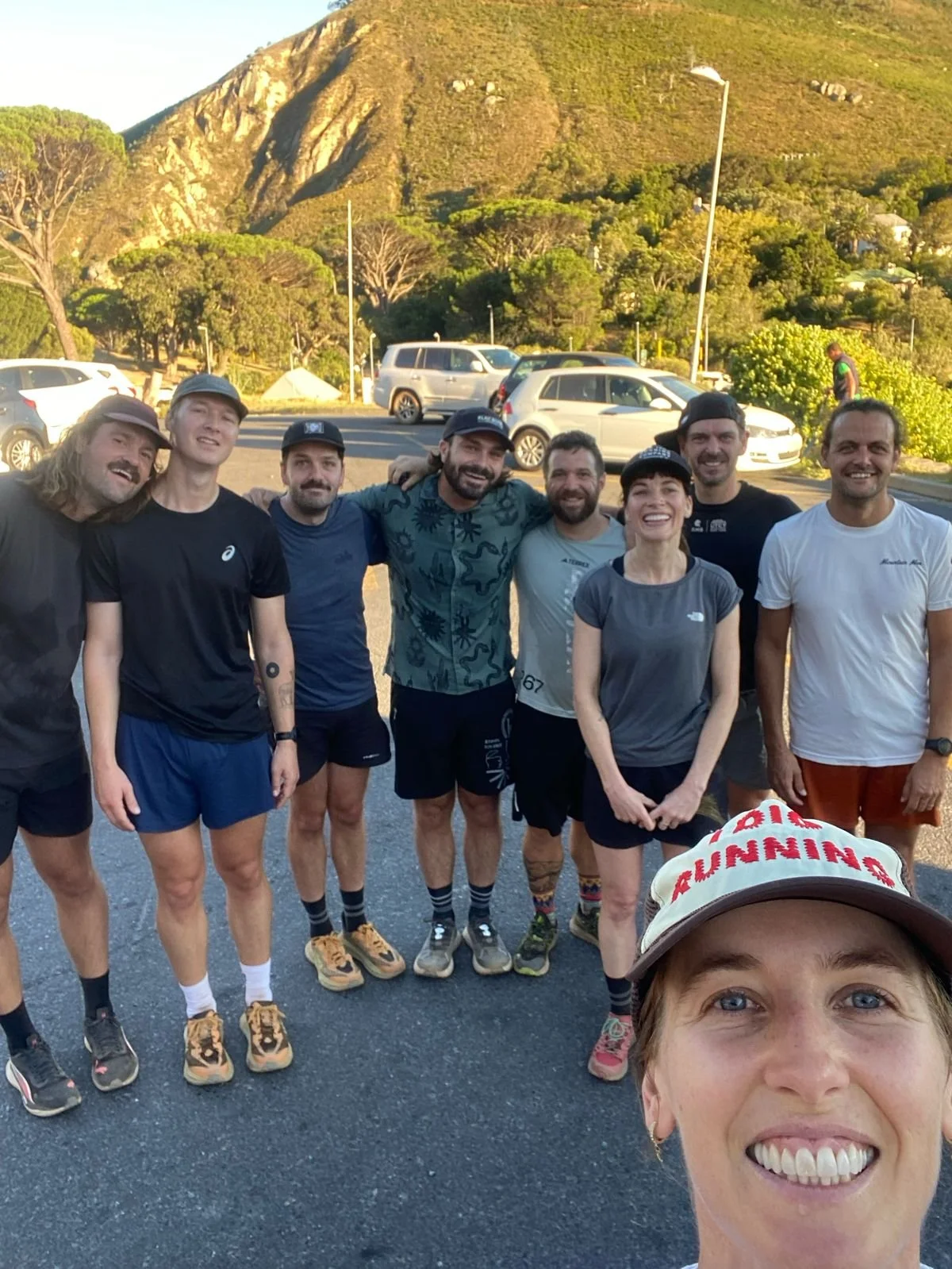 A group of nine smiling people, dressed in athletic clothing, posing outdoors in front of a mountain, trees, and parked cars during daylight.