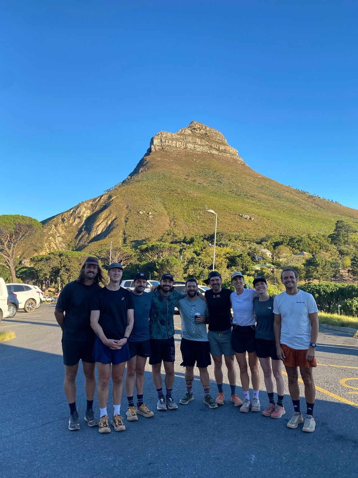 A group of nine people standing together in a parking lot with a mountain in the background, clear blue sky, and green trees.