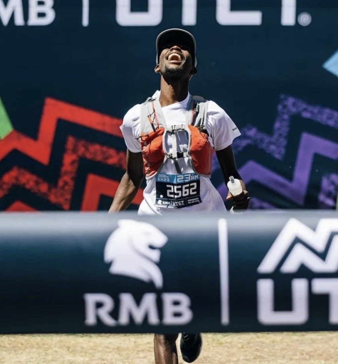 A male marathon runner crossing the finish line, smiling and wearing a white shirt, a cap, and an orange hydration pack, holding a water bottle in his right hand, with race bib number 2562.