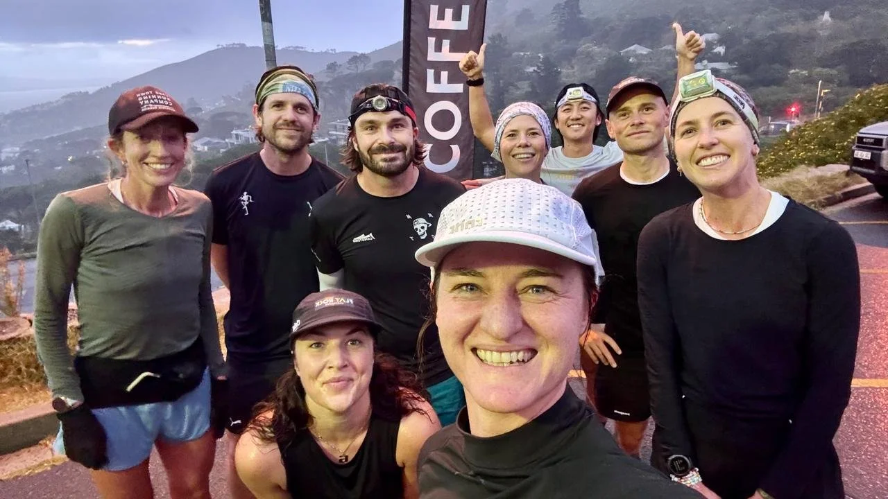 A group of nine people outdoors at dusk, dressed in athletic gear, smiling, with some wearing headlamps and a 'COFFEE' sign behind them, on a hilly road with foggy mountains in the background.