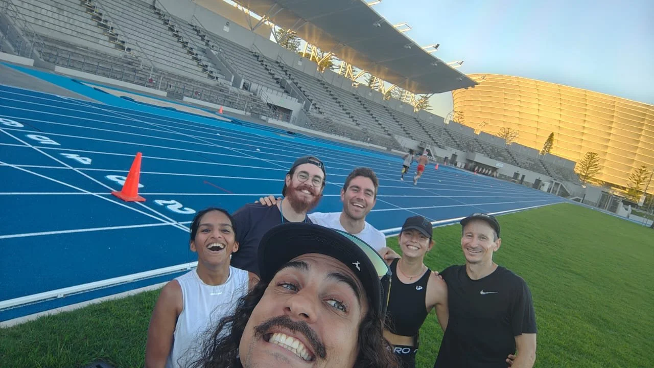A group of six people smiling for a selfie on a blue outdoor running track, with a stadium and gold-colored building in the background.